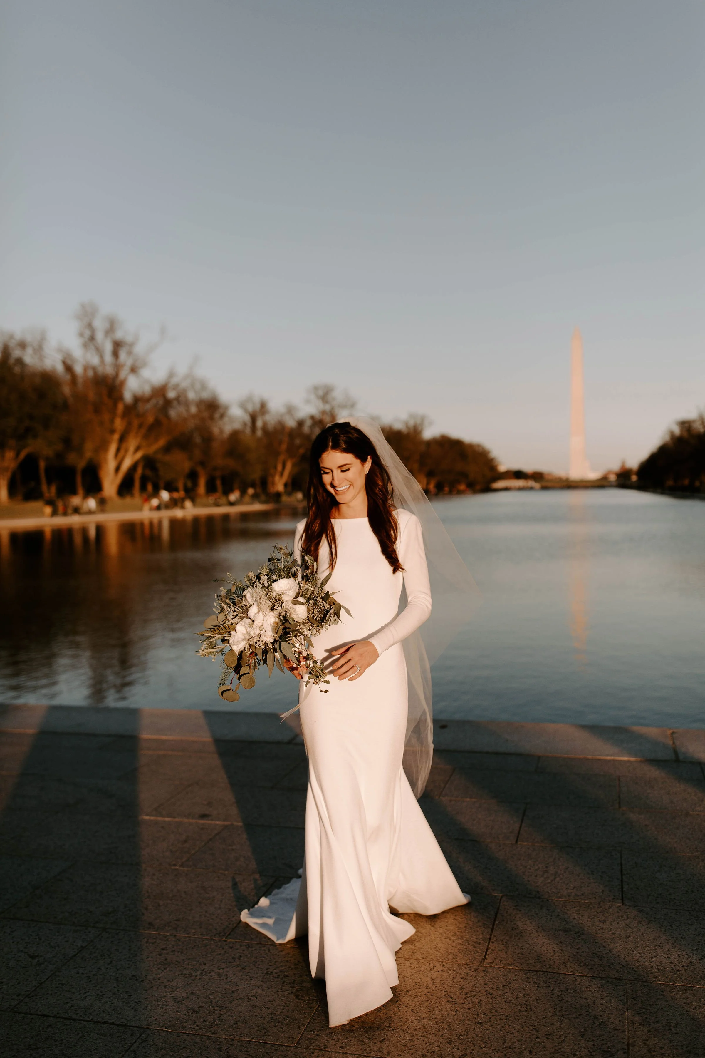 A bride in a white long-sleeve wedding gown holding a bouquet of flowers standing near the Lincoln Memorial Reflecting Pool at sunset with the Washington Monument in the background.