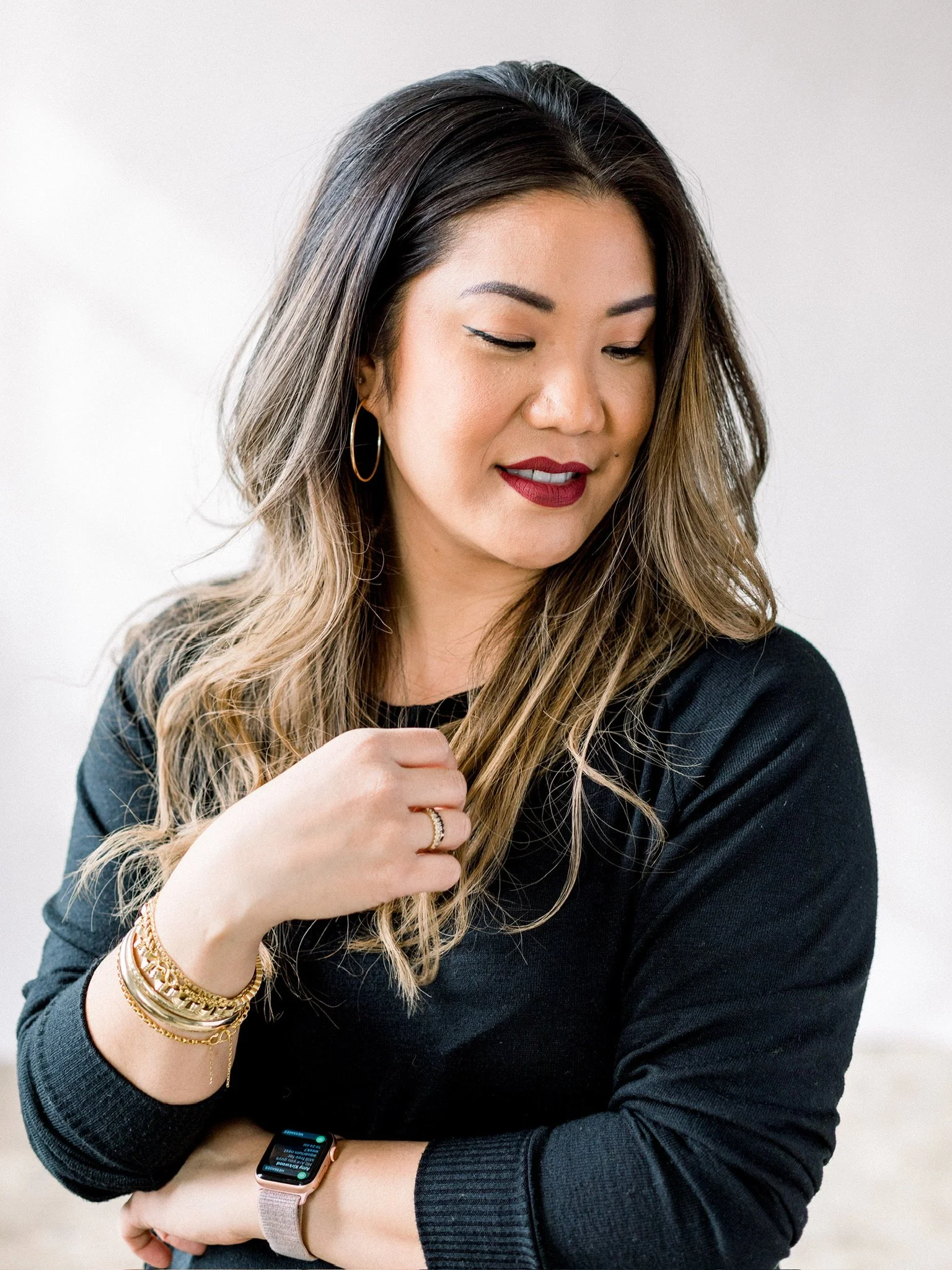 A woman with long, wavy hair, makeup, and jewelry, wearing a black top, looking down with a slight smile.