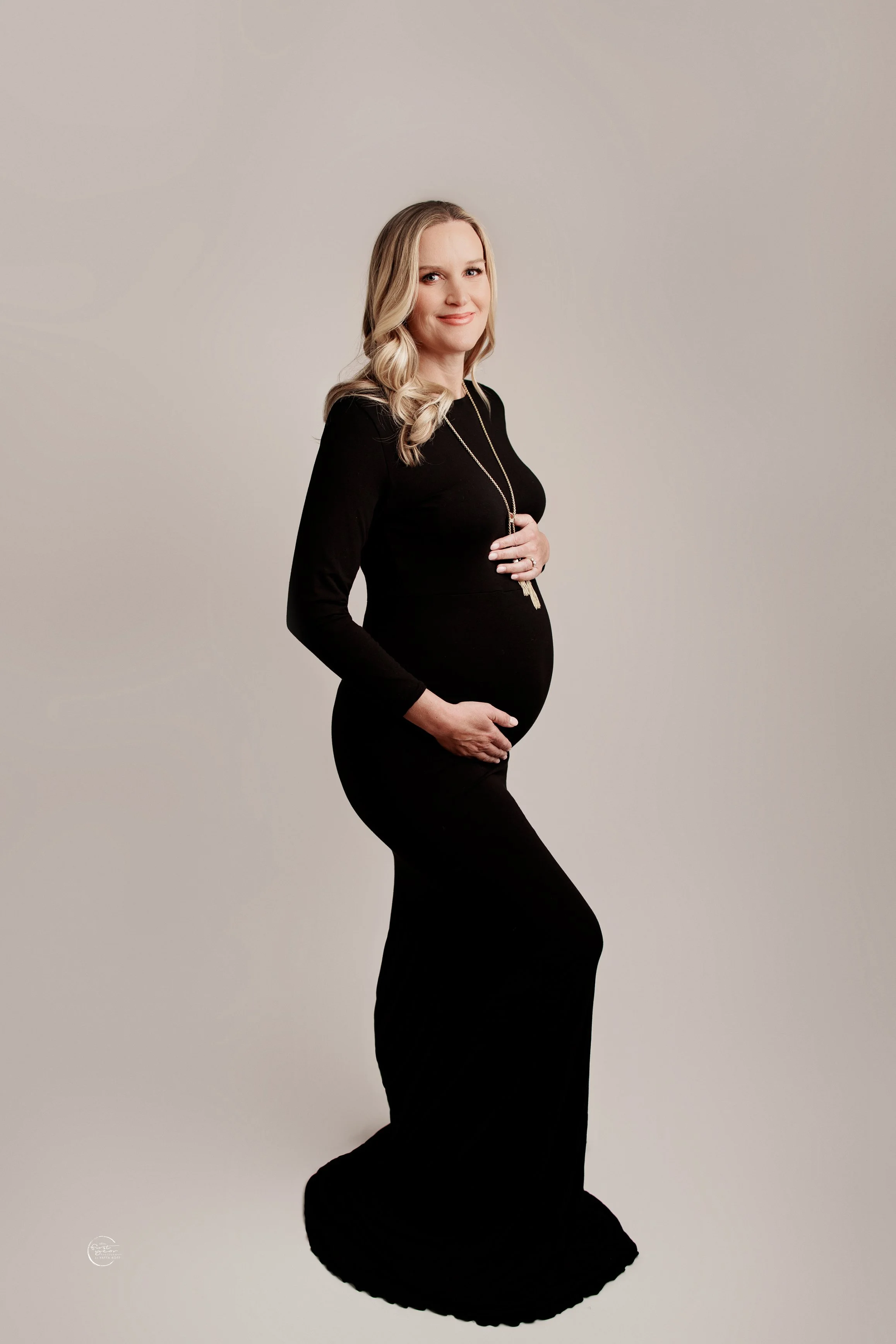 A pregnant woman standing sideways, wearing a fitted black dress, smiling softly, with her right hand on her belly and her left hand gently holding her belly, against a neutral background.