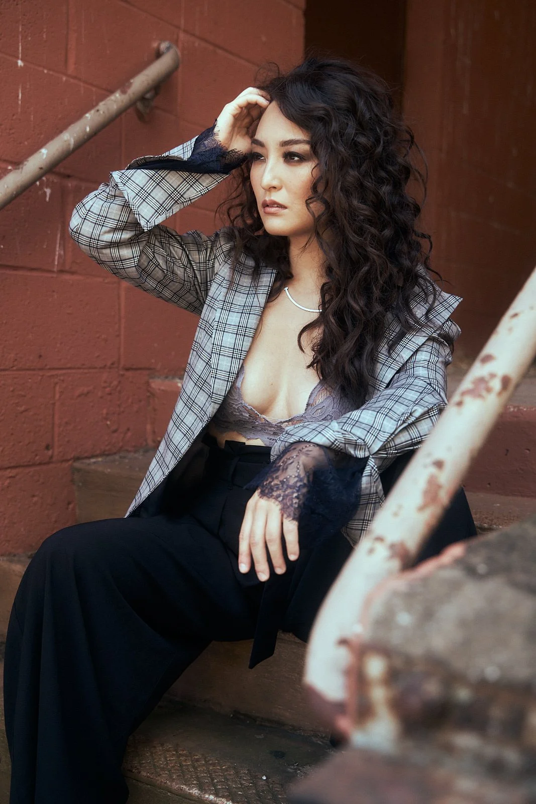 A woman with long curly dark hair sitting on outdoor stairs, wearing a plaid blazer with lace cuffs, a lace bralette, and black high-waisted pants, against a red brick wall background.
