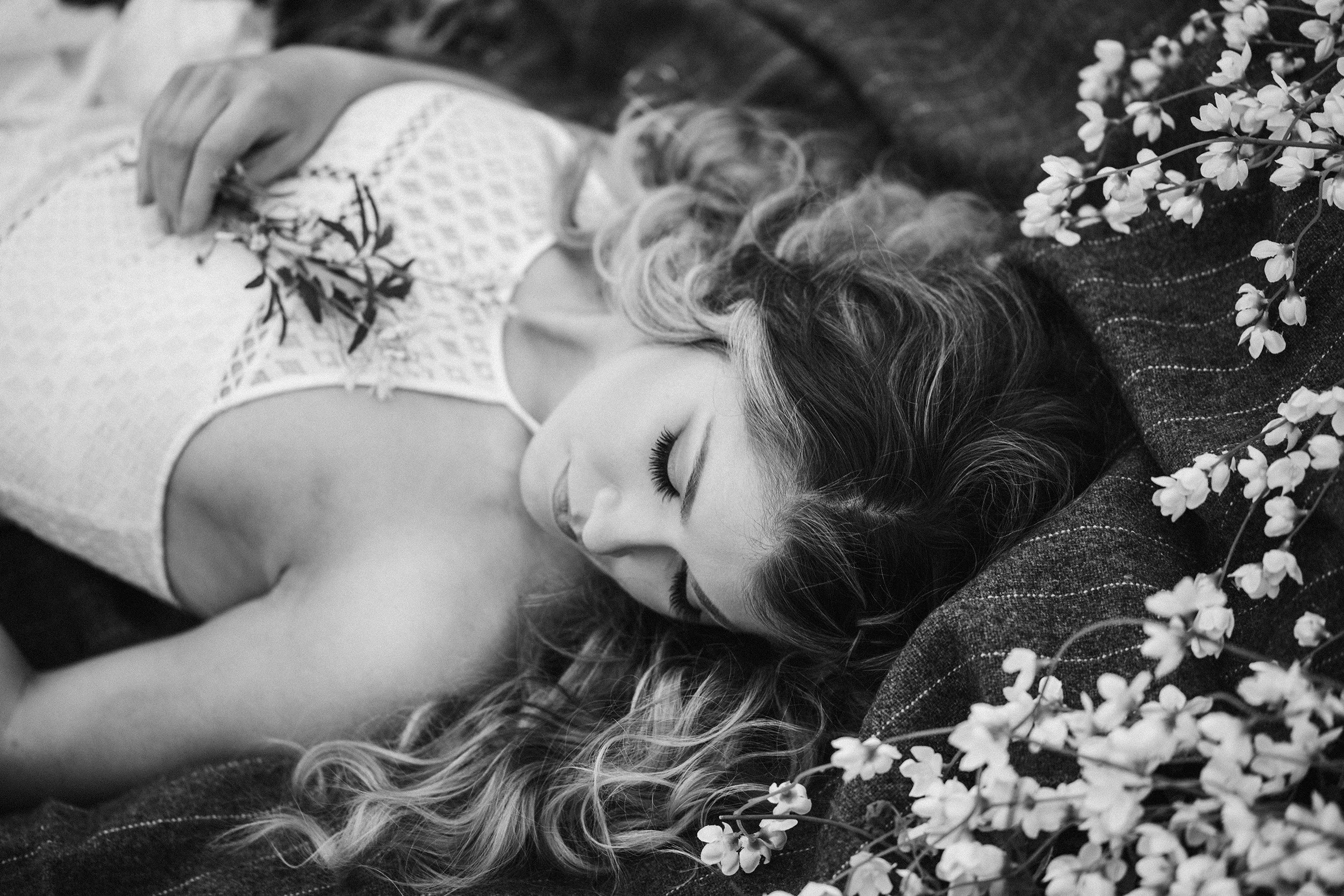 Black and white photo of a woman lying on a dark textured surface with her eyes closed. She has long curly hair spread around her head and is wearing a sleeveless, patterned top. Flowers are arranged near her head.