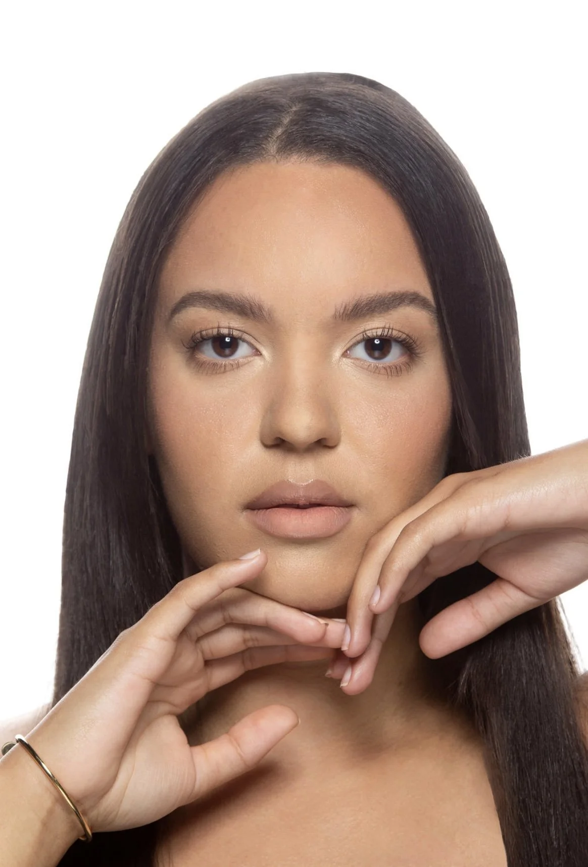 Close-up of a young woman with long dark hair, neutral makeup, and a neutral expression, holding her hand near her face against a white background.