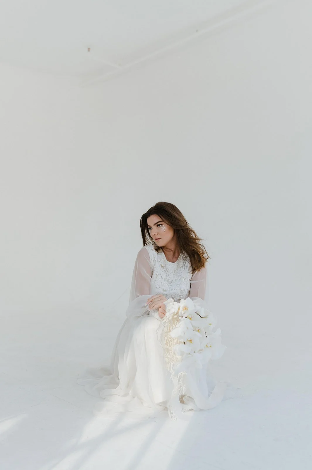 A woman in a white dress sitting on the floor holding a bouquet of white flowers in a minimalistic, white studio setting.