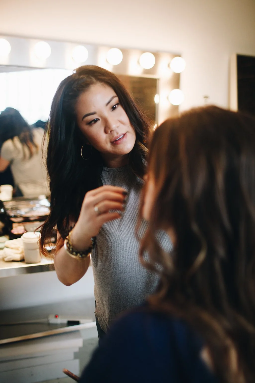 A woman with long dark hair and hoop earrings is applying makeup or grooming a person with medium-length brown hair, in a well-lit room with vanity mirrors and round lights.