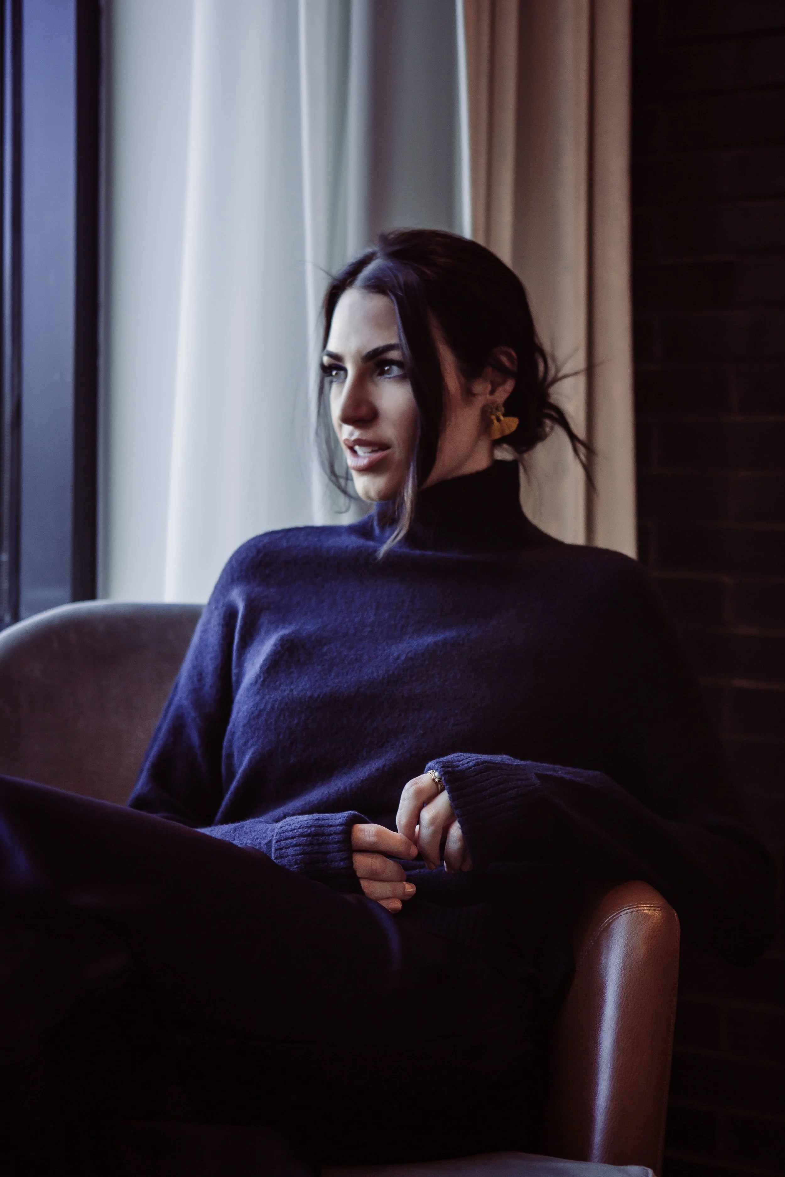 Woman with dark hair, wearing a navy blue sweater, sitting by a window, looking thoughtful.
