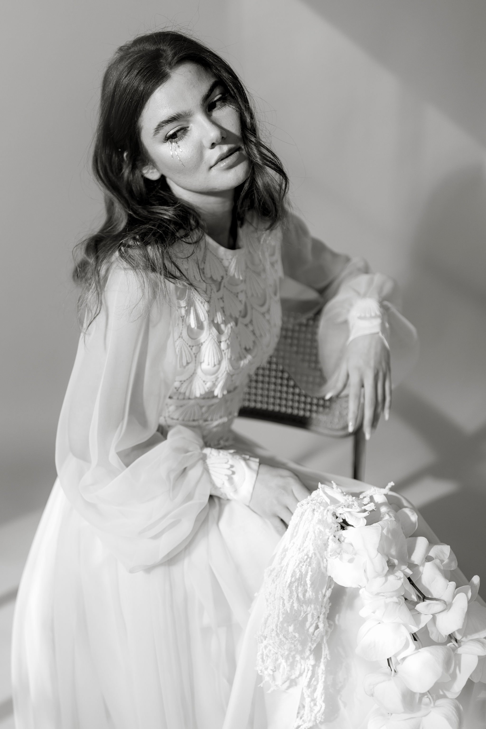 A young woman with wavy hair and tears on her cheek, sitting on a chair in a wedding dress, holding a bouquet of flowers.