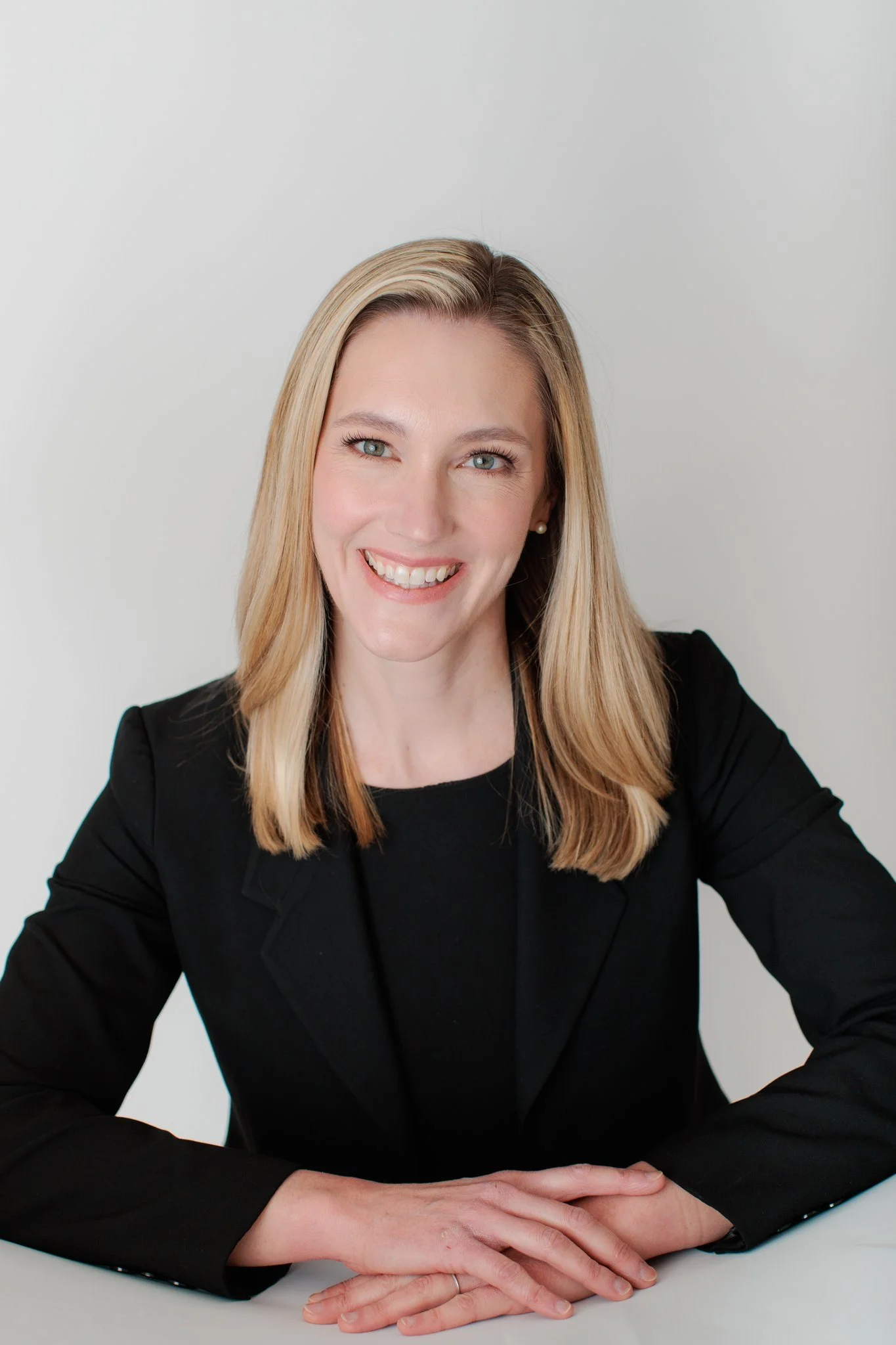A woman with blonde hair, blue eyes, and a big smile, wearing a black blazer and sitting at a white table with her hands resting on it, against a plain light gray background.