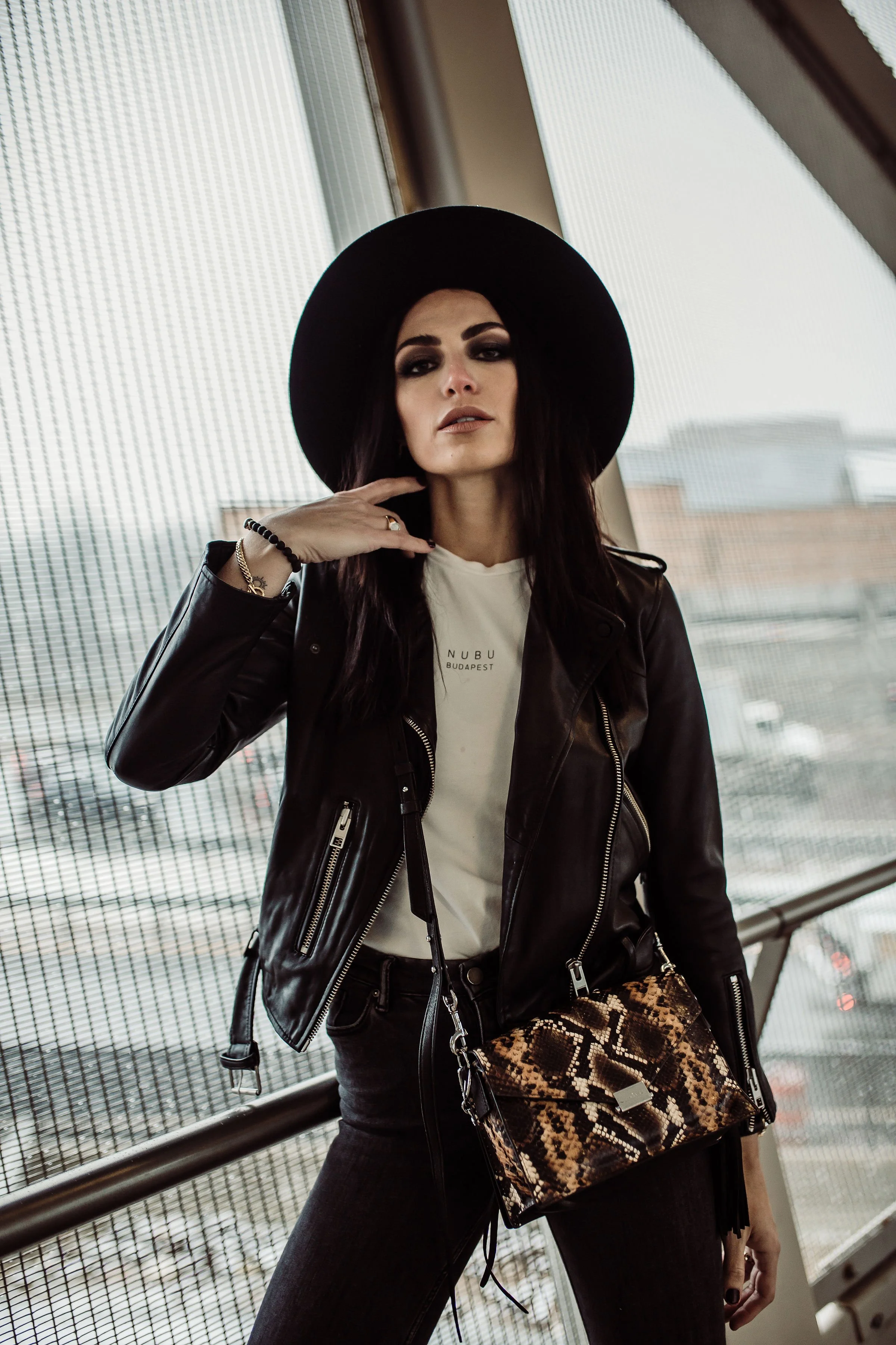 A woman with dark hair and makeup wearing a black wide-brim hat, black leather jacket, white T-shirt, black pants, holding a snake-skin patterned purse, standing indoors near large windows.