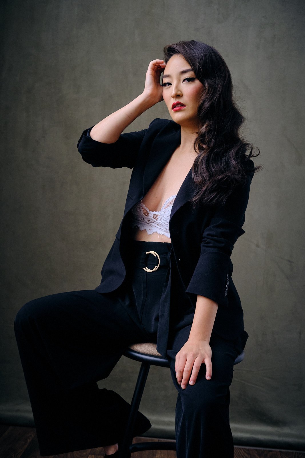A woman with wavy black hair poses sitting on a stool against a plain background, wearing a black blazer, black pants with a gold belt, and a white lace crop top.