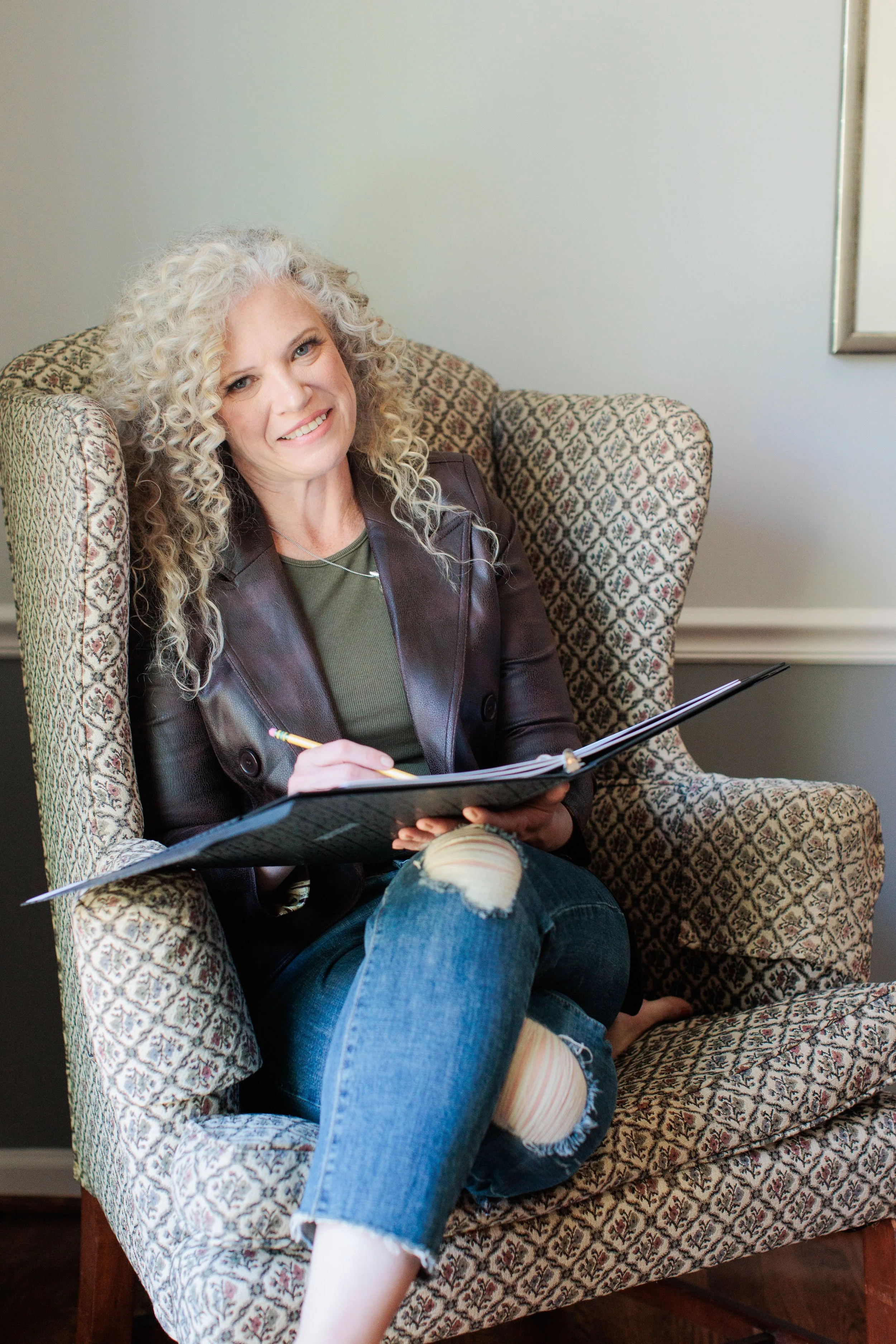 A woman with curly blonde hair smiling while sitting in an armchair, holding a notebook and pen.