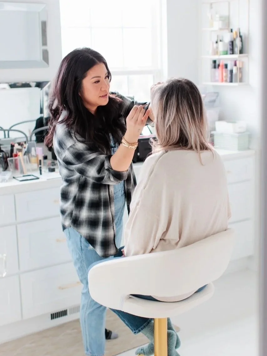 Makeup artist applying makeup to woman sitting in a white salon chair in a brightly lit room.
