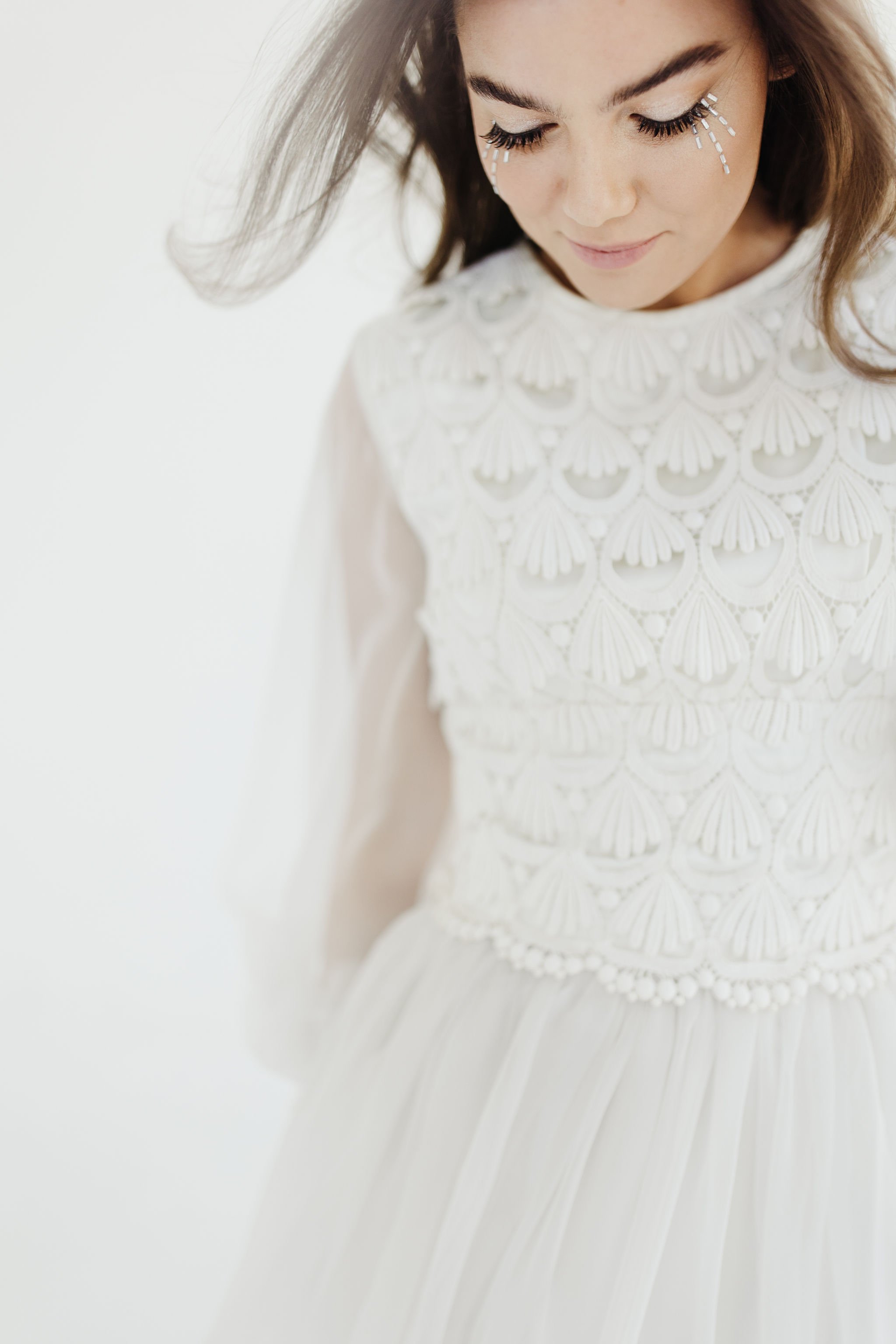 Woman with face paint, wearing a white dress with ornate embroidery and sheer sleeves, looking down with a gentle smile.