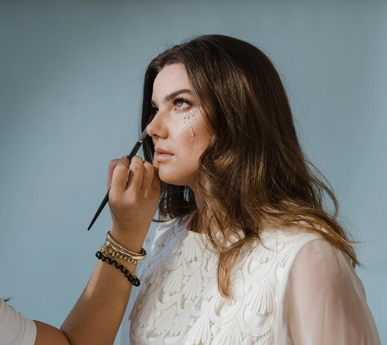 A woman with brown wavy hair is having makeup applied to her face by a makeup artist. The artist is using a brush near her right eye, and there are white dotted lines drawn under her eye, suggesting she's getting ready for a special look or photo shoot.