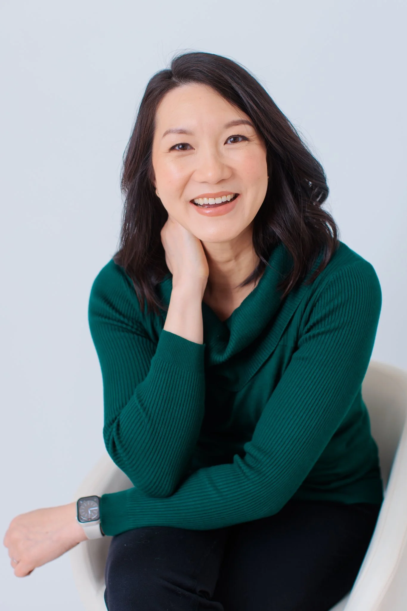 Portrait of a smiling woman with shoulder-length dark hair wearing a green long-sleeve top, sitting on a white chair against a light background.