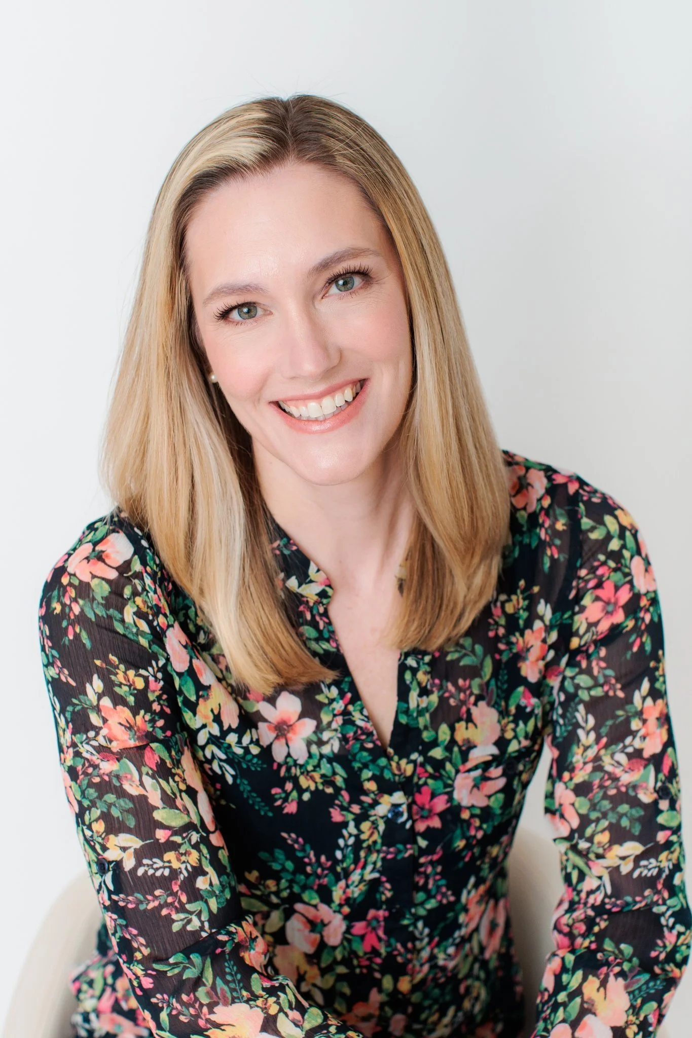 A woman with blonde hair smiling, wearing a black floral blouse, against a light background.