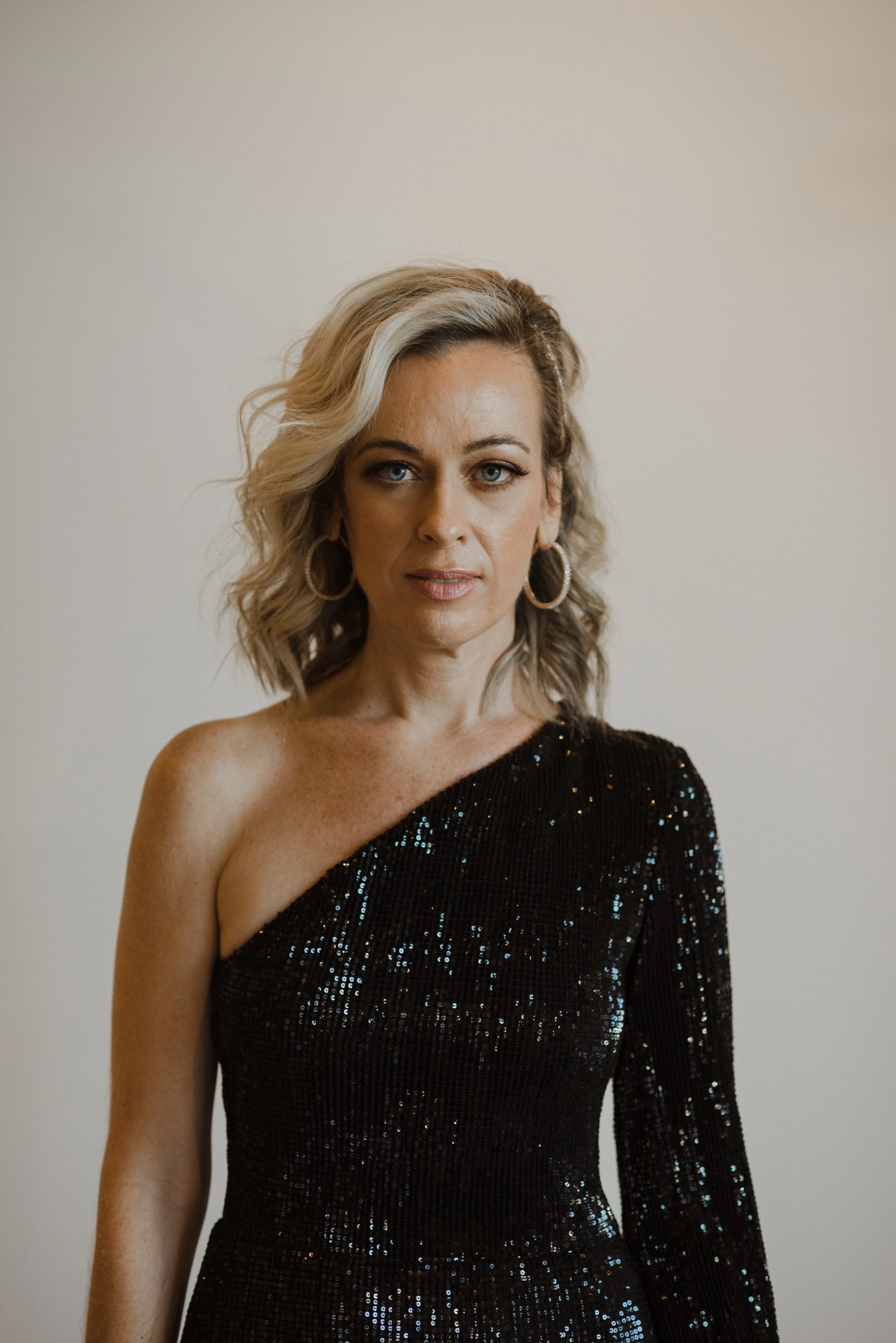 A woman with wavy blonde hair, wearing a one-shoulder black sequined dress.