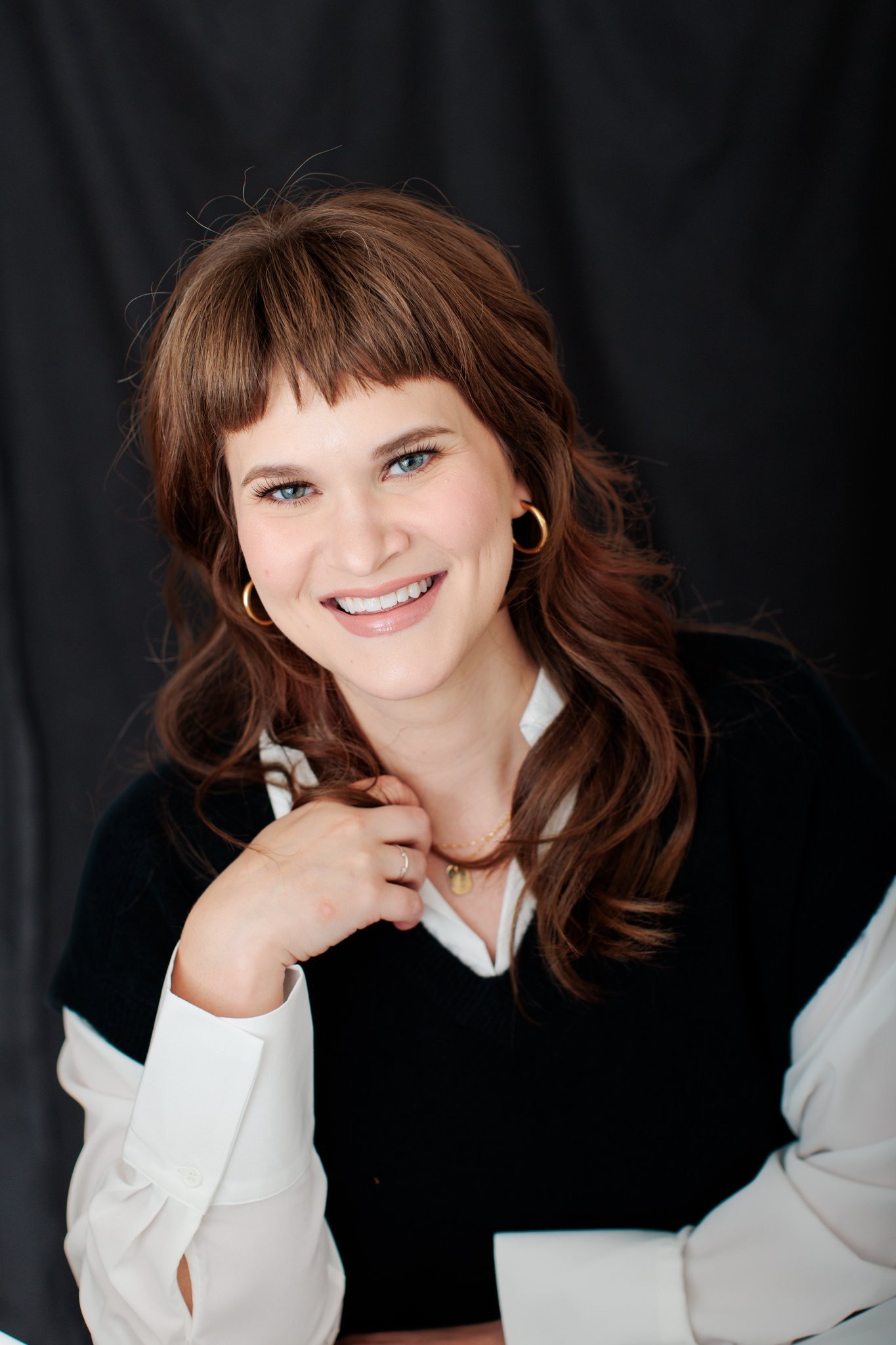 Portrait of a woman with shoulder-length auburn hair, blue eyes, wearing gold hoop earrings, a black and white collared shirt, and gold jewelry, smiling and resting her chin on her hand against a dark background.