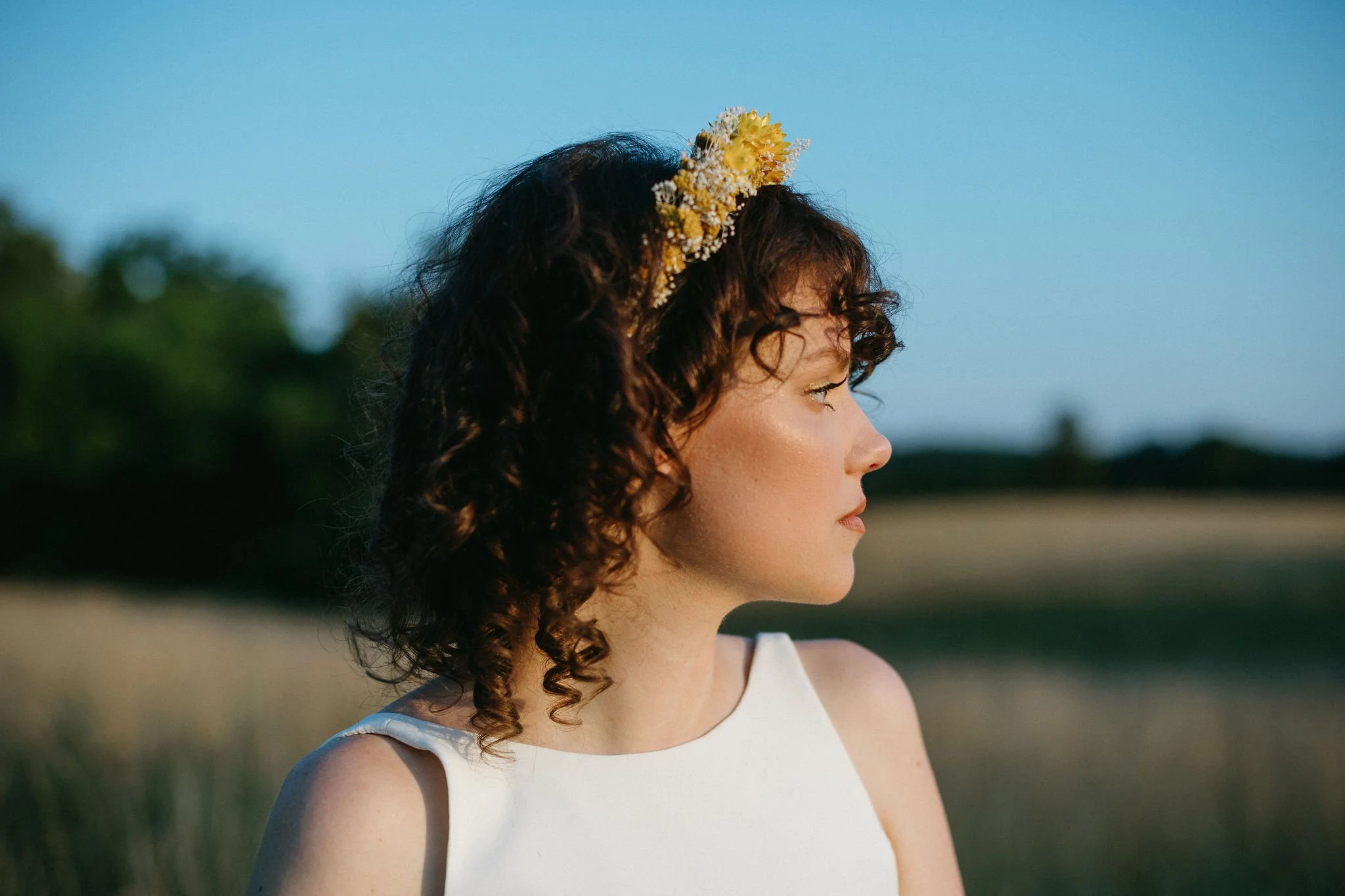 A woman with dark, curly hair wearing a flower crown looking to the right outdoors during sunset.