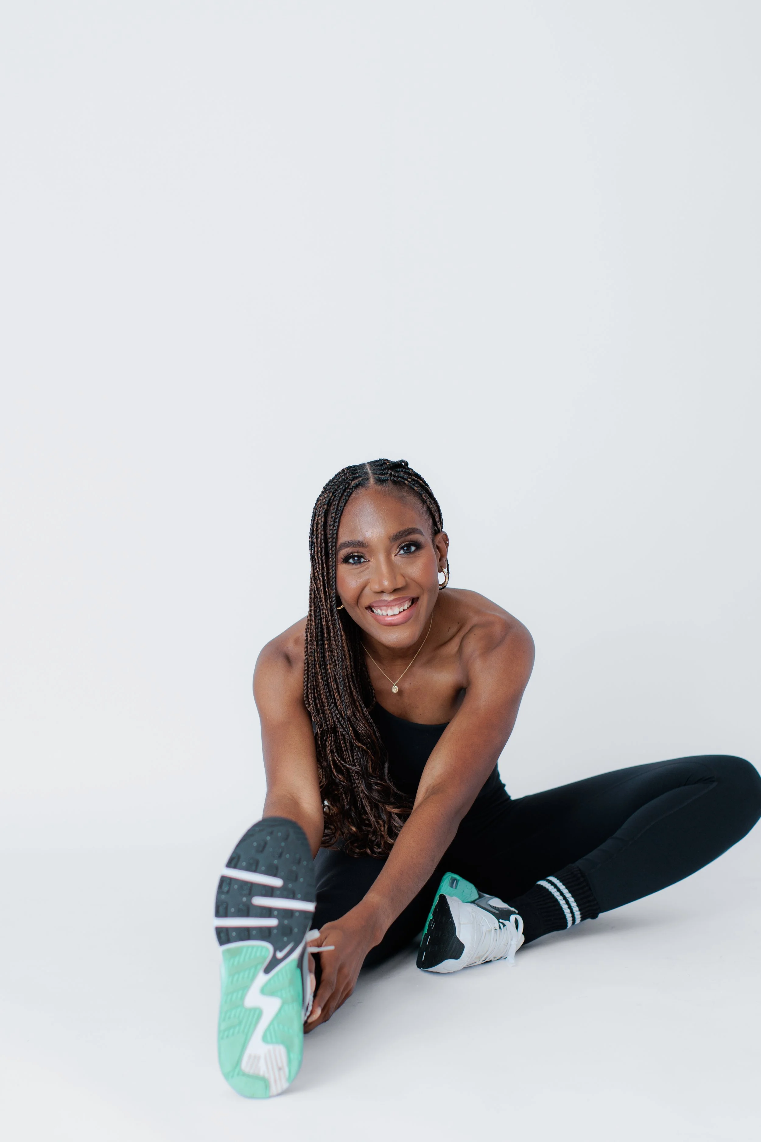 A woman with braided hair, wearing a black tank top and black leggings, sits on the floor smiling, holding her left foot in front of her with a athletic shoe, against a plain white background.