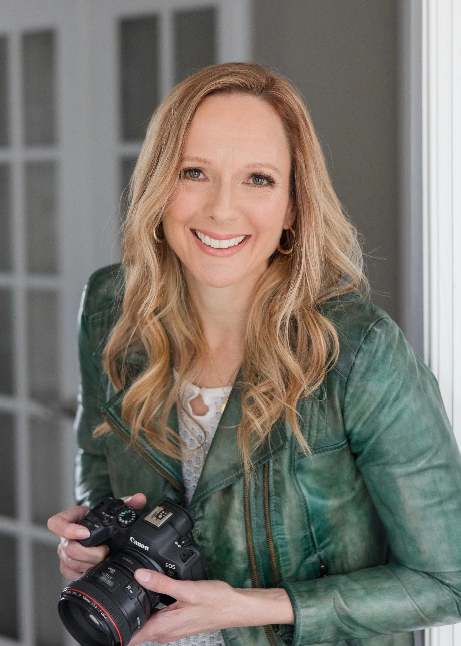 A woman with long blonde hair holding a Canon camera, smiling, wearing a green jacket and a white lace top, standing indoors near a window.