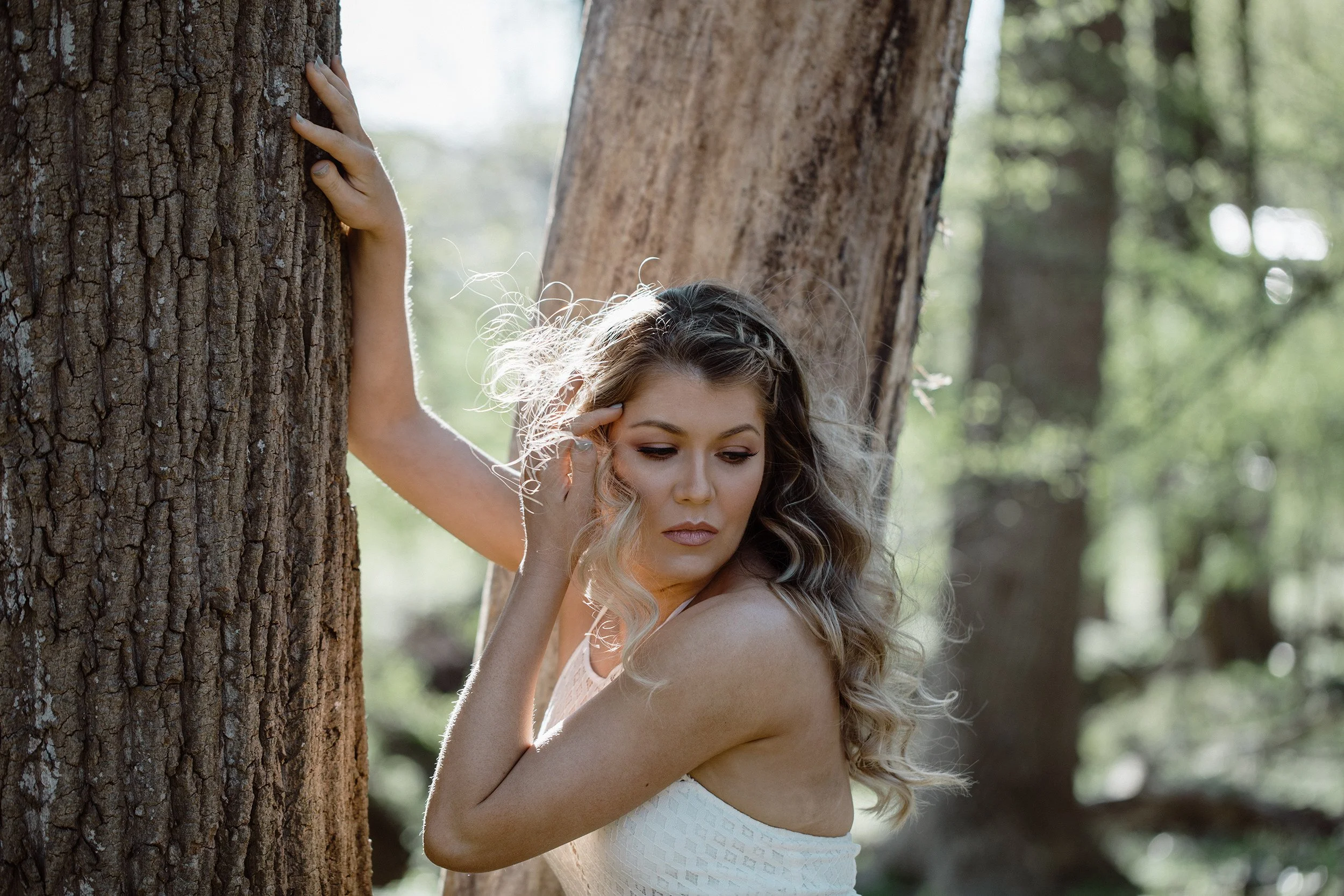 A woman with wavy hair leaning against a tree in a forest with sunlight filtering through the leaves.