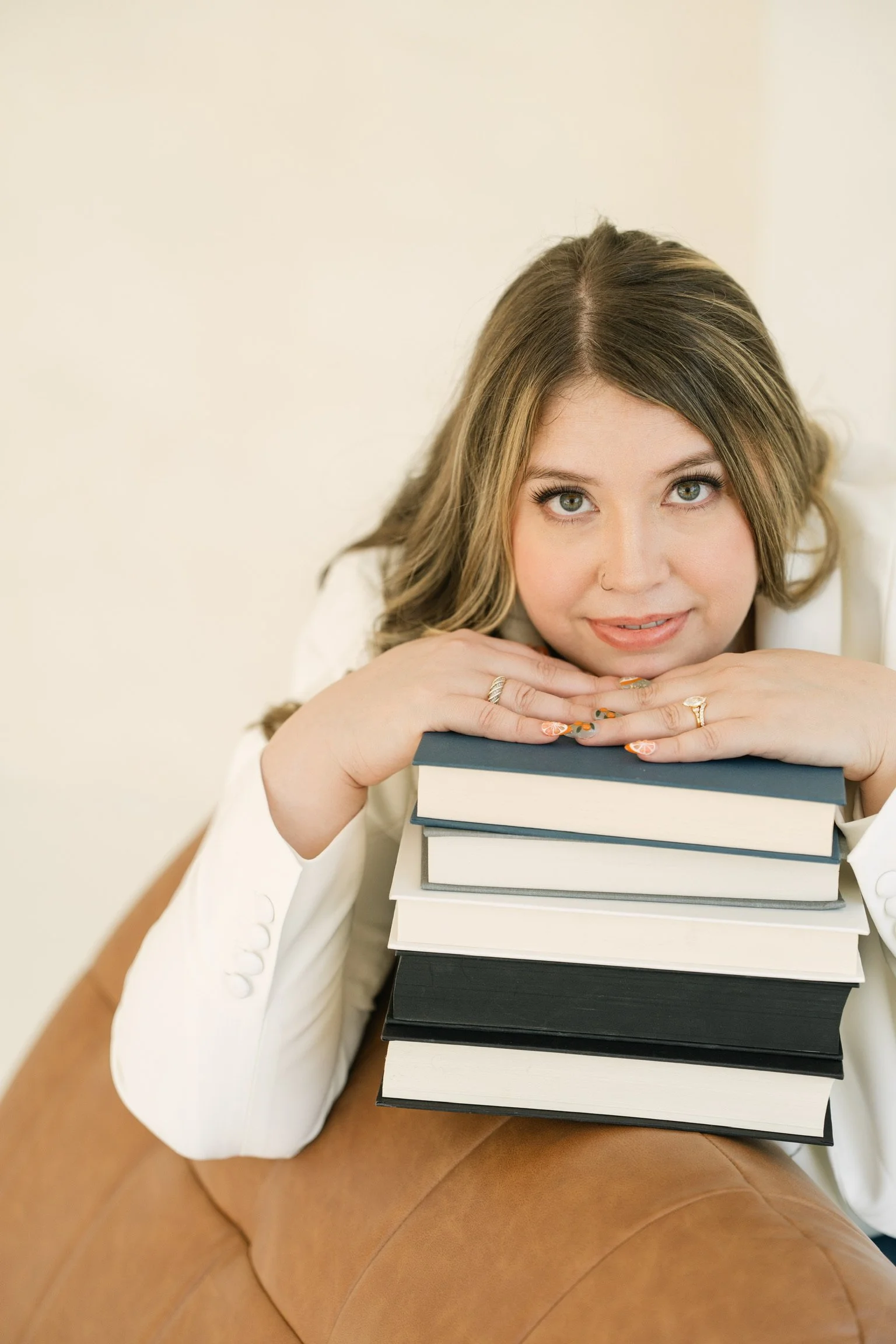Young woman with brown hair and light eyes resting her face on a stack of books, looking at the camera with a gentle expression, sitting on a tan leather couch against a plain off-white background.