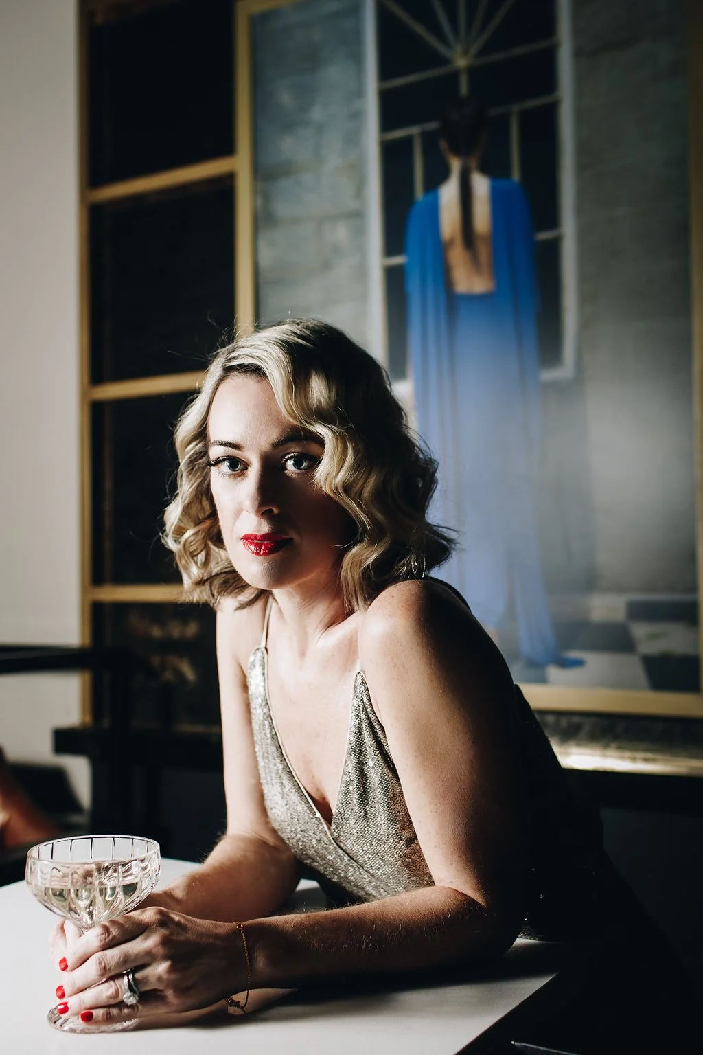 A woman with styled blonde hair and red lipstick, wearing a glittery sleeveless dress, sitting at a table in a dimly lit room with a painting of a woman in a blue dress hanging on the wall behind her.