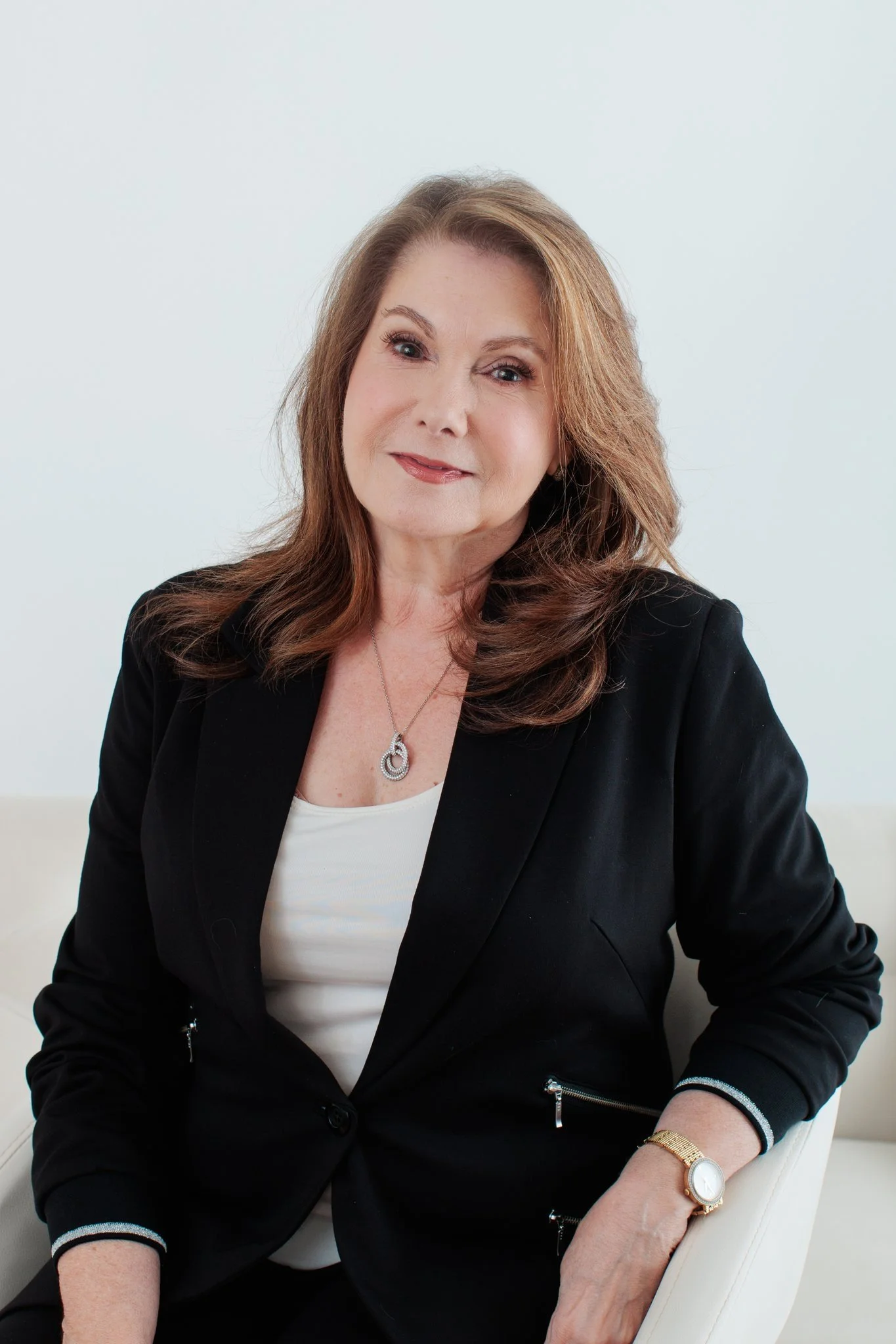 A middle-aged woman with wavy, reddish-brown hair wearing a black blazer, white top, gold watch, and silver jewelry, sitting on a light-colored sofa against a white background.