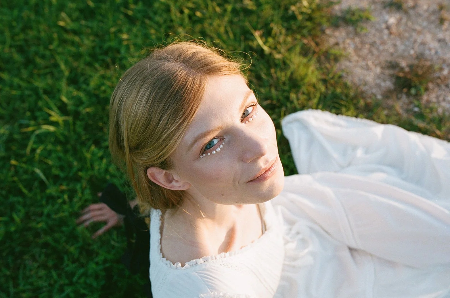 A young woman with blonde hair and blue eyes lying on grass, wearing a white dress with pearl accents near her eyes, captured from above during golden hour.