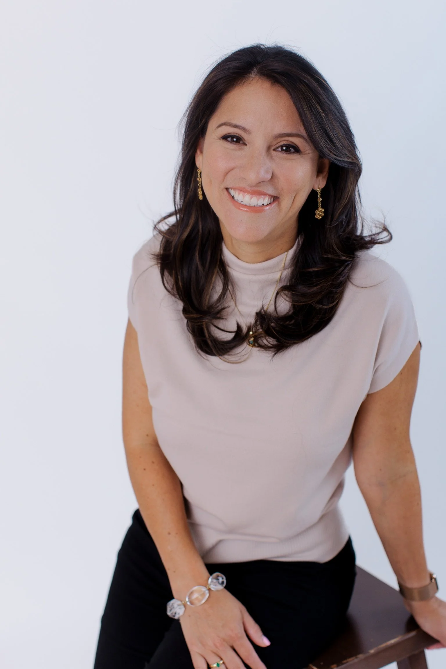 A woman with shoulder-length dark wavy hair, smiling, wearing a light-colored top, black pants, gold earrings, and a bracelet, leaning on a chair against a plain white background.