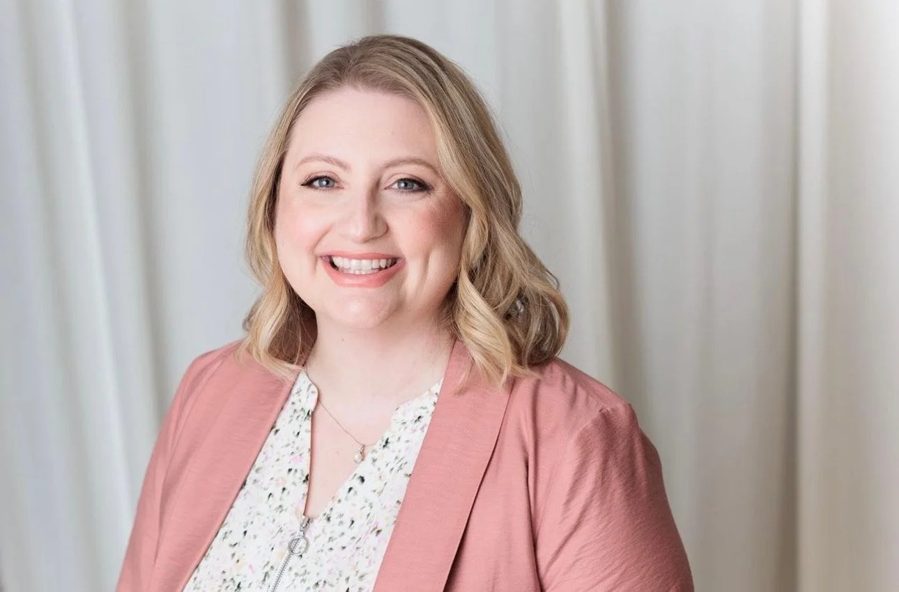 Portrait of a smiling woman with blonde hair, wearing a pink blazer and a white blouse, standing in front of a beige curtain.