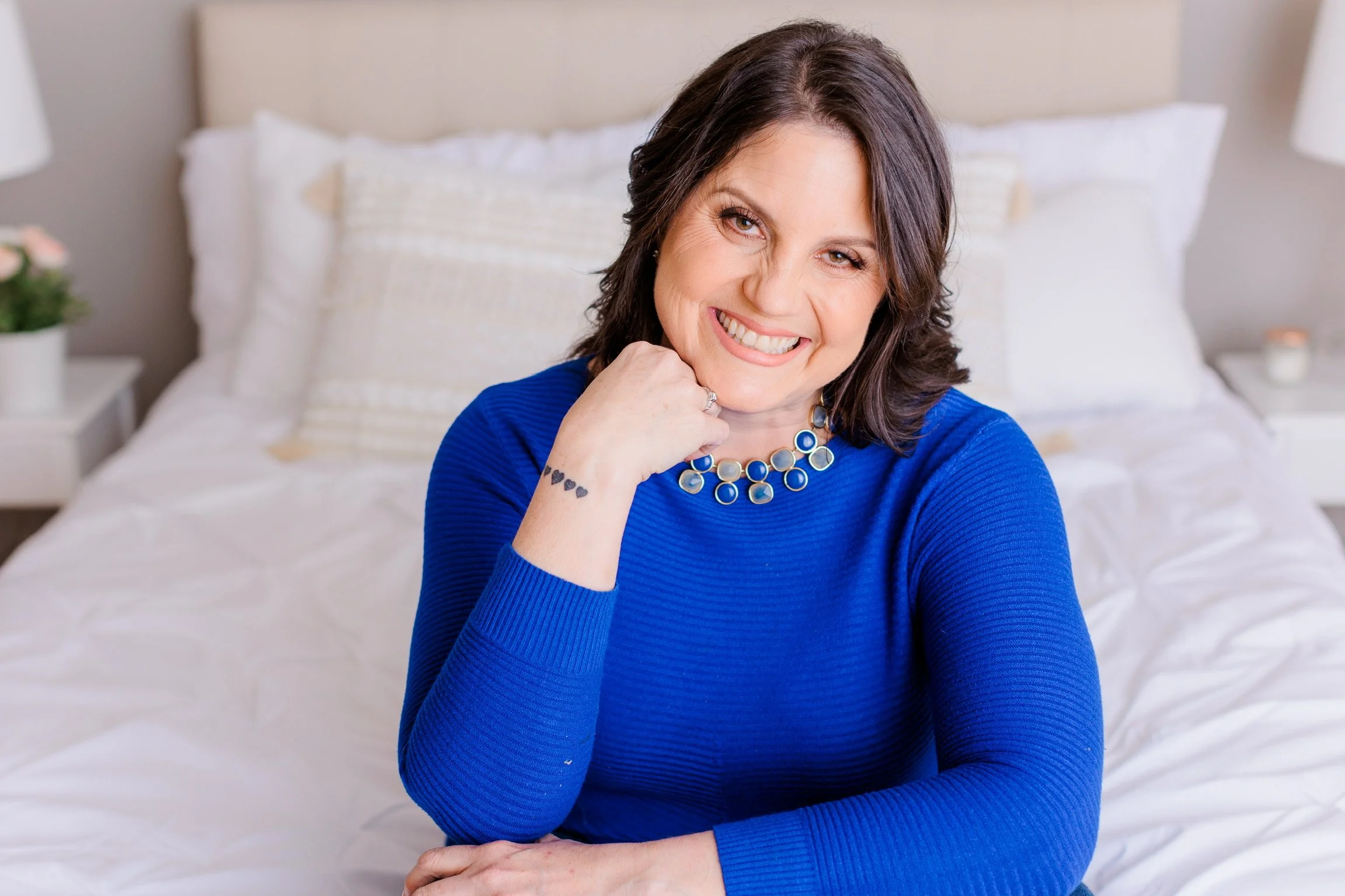 Woman with dark brown hair in a blue long-sleeve shirt and statement necklace sitting on a bed, smiling at the camera.
