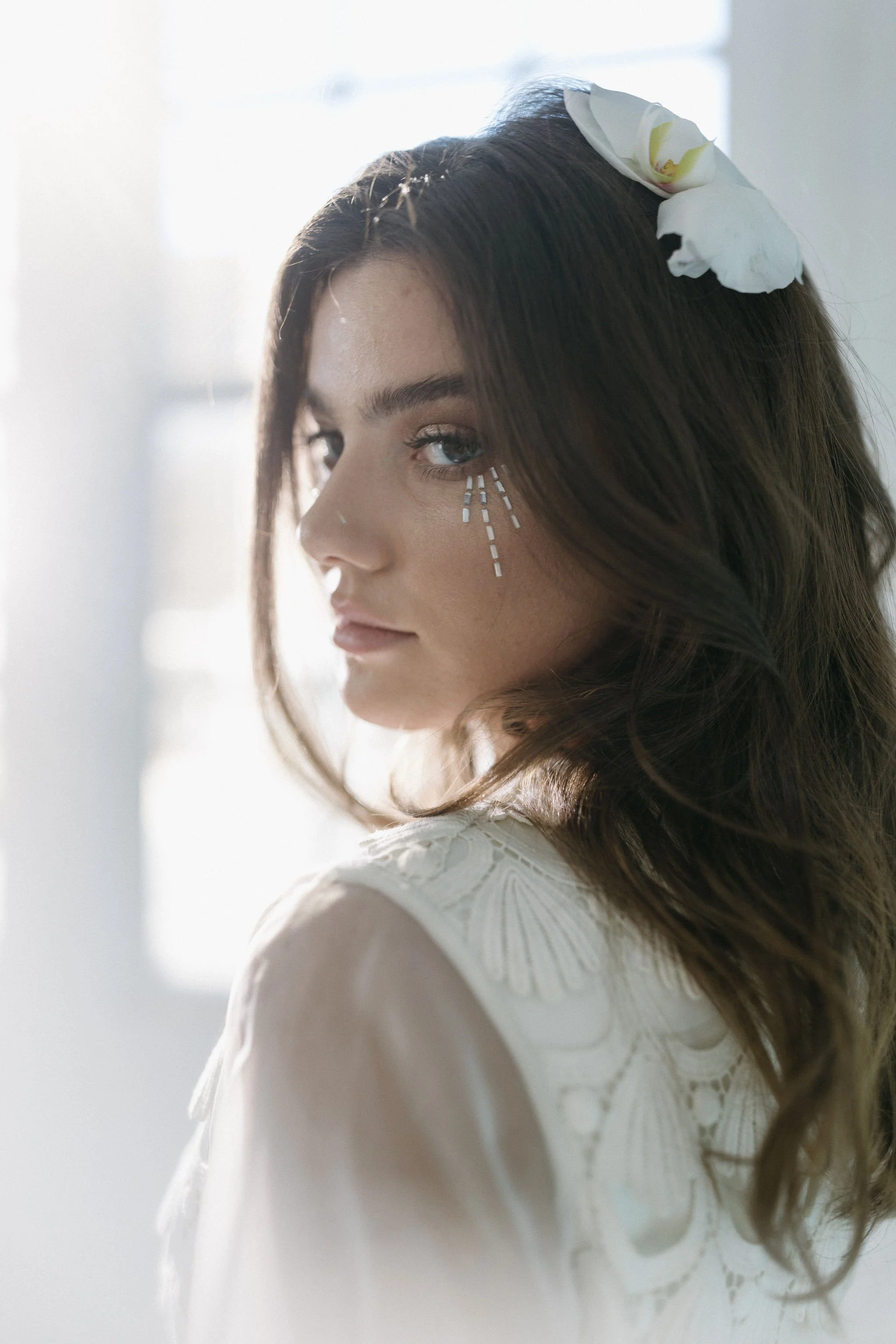 A young woman with blue eyes and brown hair, wearing a white dress with embroidered details, has a white flower in her hair and small white decorative lines under her eye.