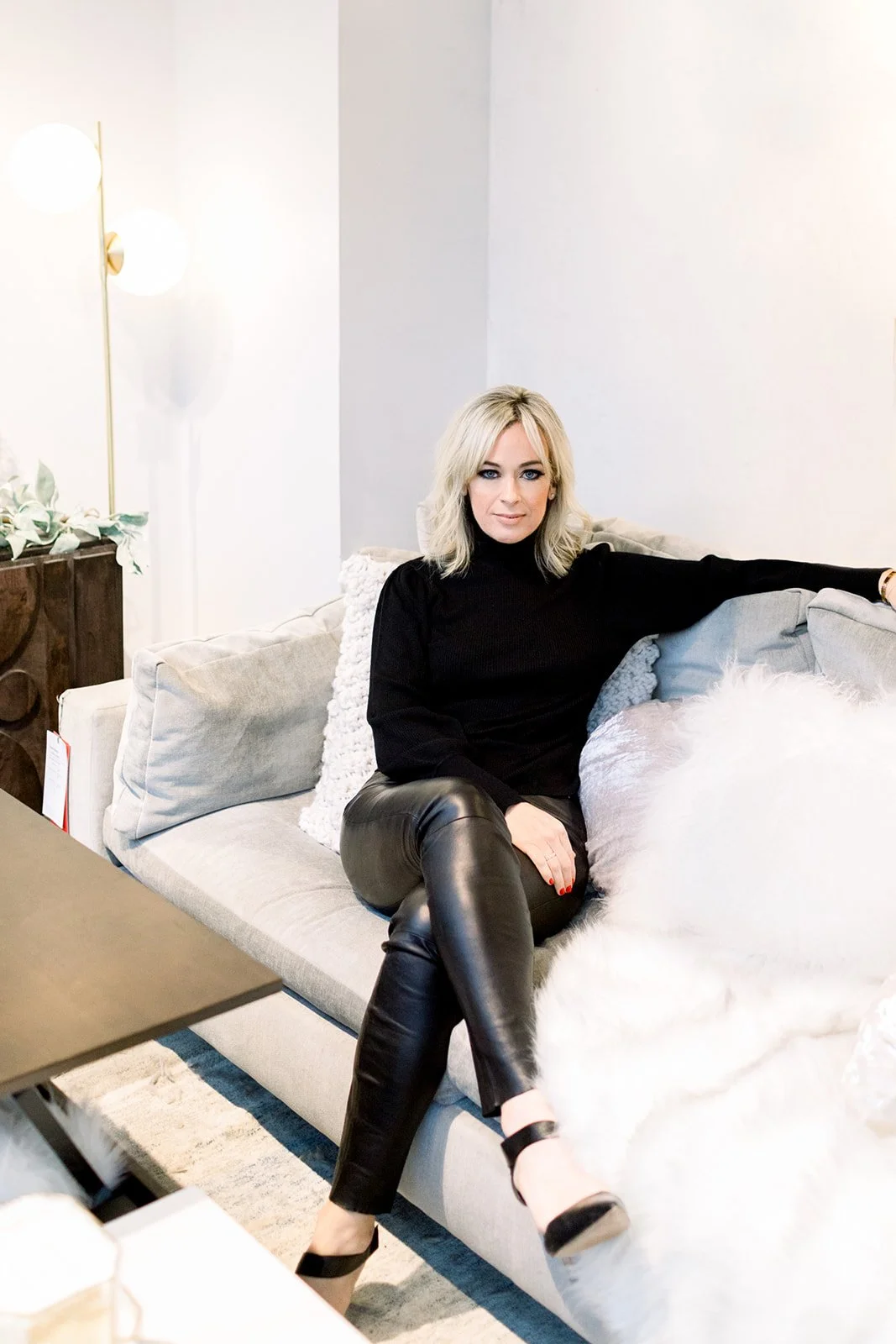 A woman with blonde hair wearing a black turtleneck and black leather pants, sitting on a light-colored sofa with pillows, in a modern, well-lit living room.