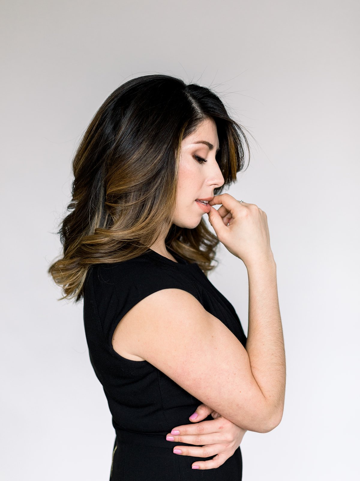 Side profile of a woman with wavy brown and blonde hair, wearing a black sleeveless top, standing against a plain white background.
