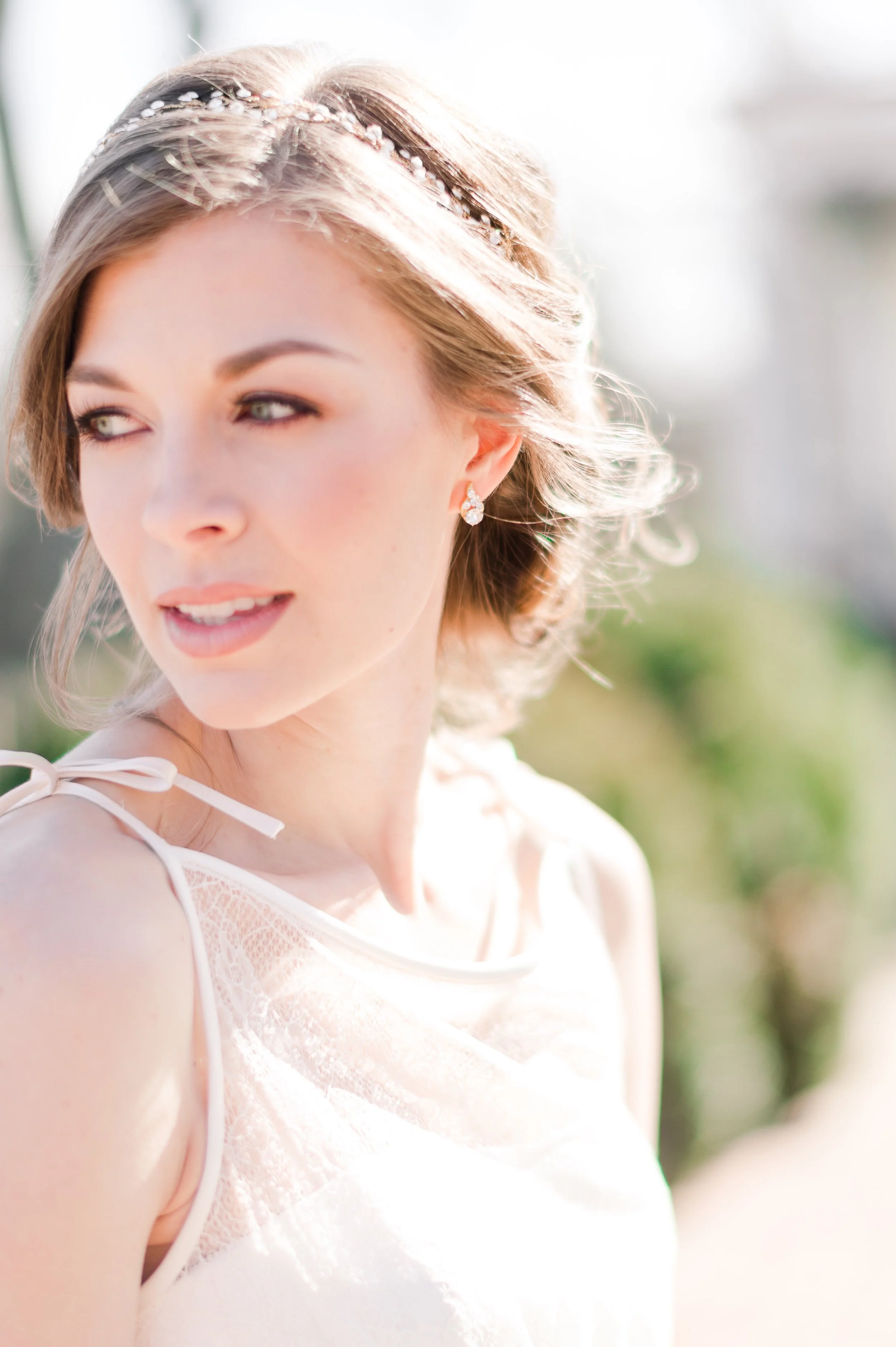 Close-up of a young woman with short brown hair, wearing a white dress with lace details and a decorative headband, outdoors during daytime.