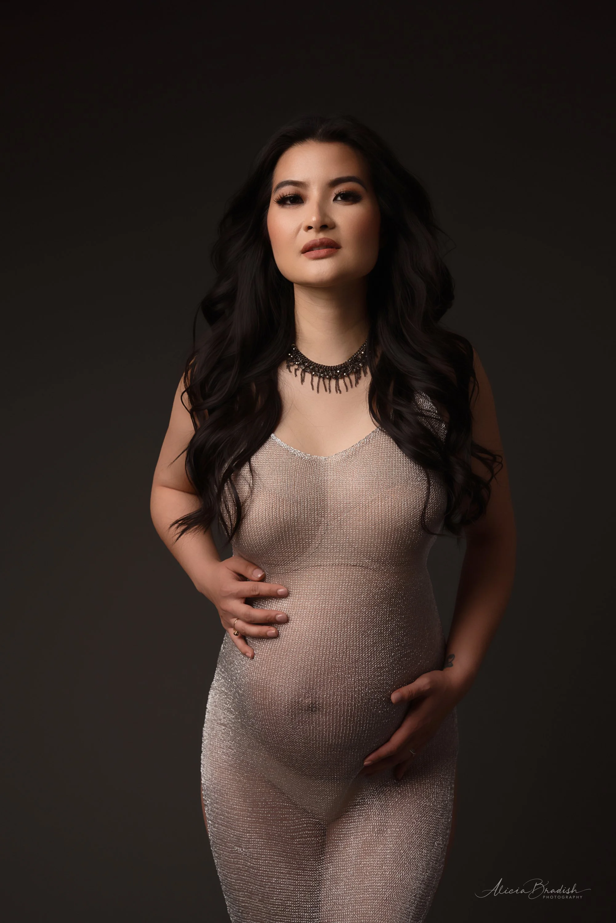 A pregnant woman with long dark hair, wearing a shimmering silver sleeveless dress and a black necklace, standing against a dark background.