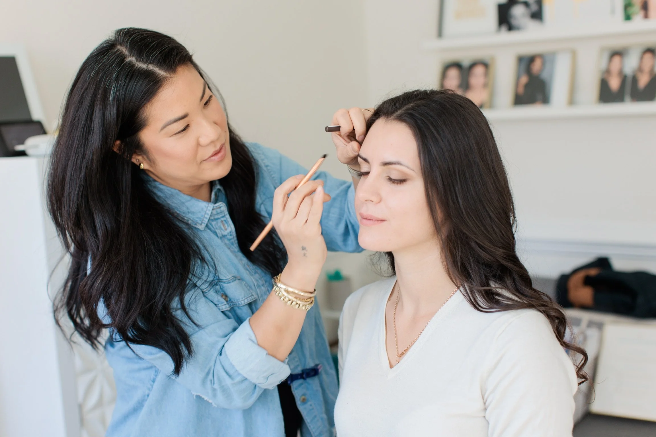 Makeup artist applying makeup to a woman in a white shirt with long dark hair, in a room with a photo display on the wall.
