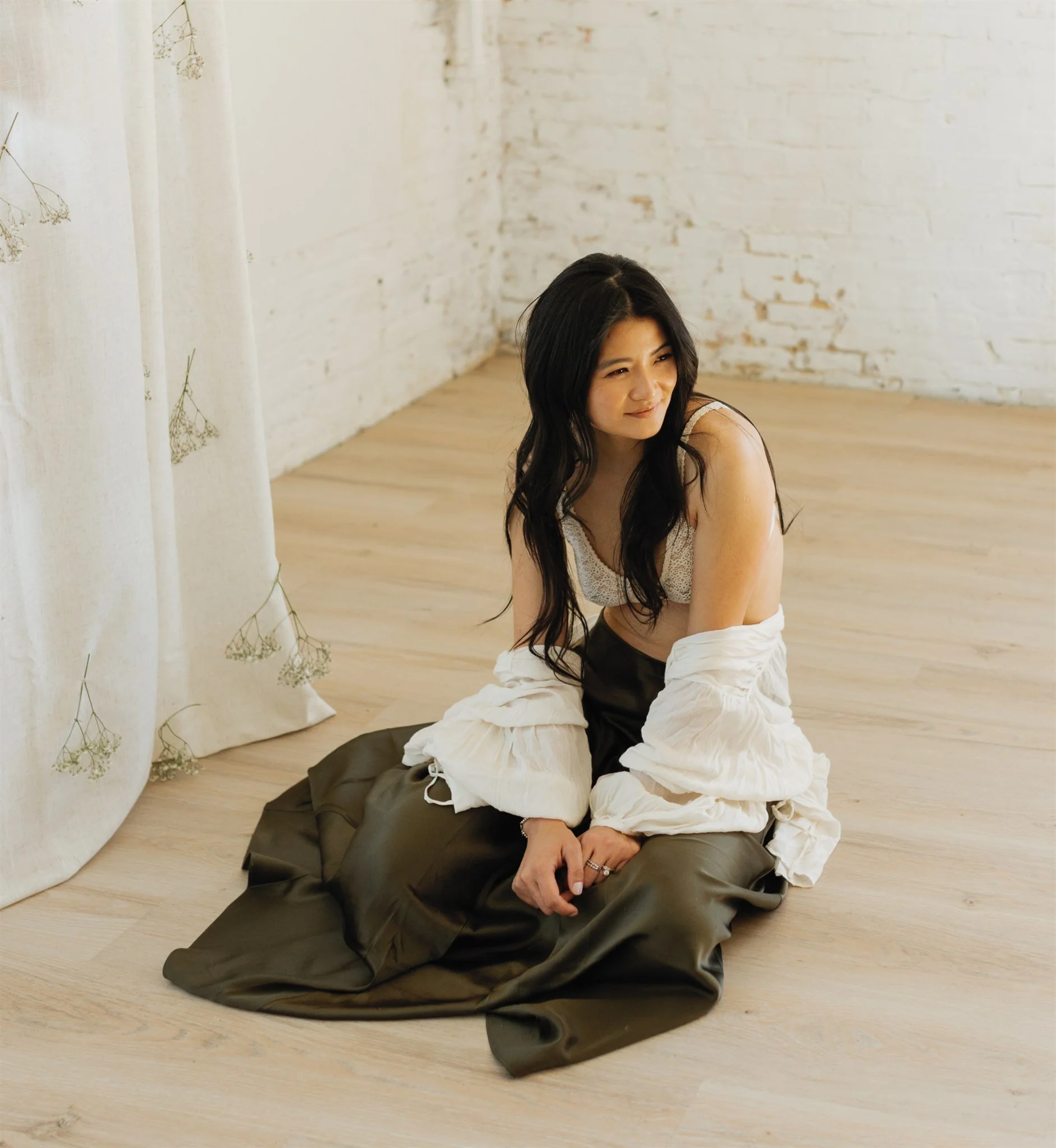 A young woman with long black hair sitting on a light wooden floor, wearing a white top and a long dark skirt, in a room with white brick walls and a white curtain with floral embroidery.