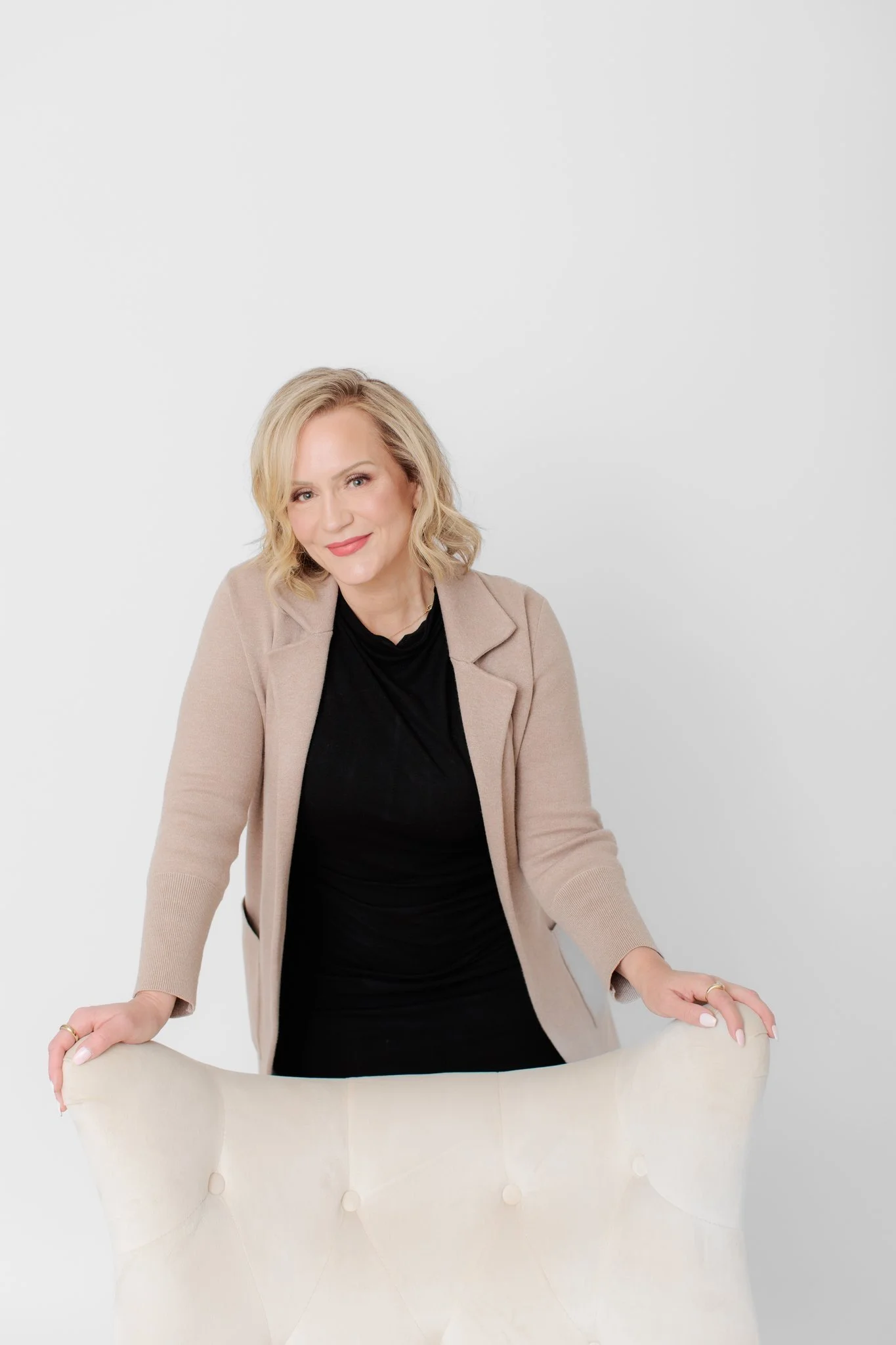 A woman with blonde hair in a beige blazer and black top, smiling, leaning on a cream-colored chair against a plain white background.