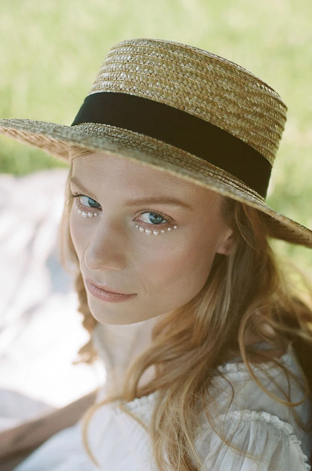 A young woman with red hair wearing a wide-brimmed straw hat with a black band, and decorative pearls under her eyes.