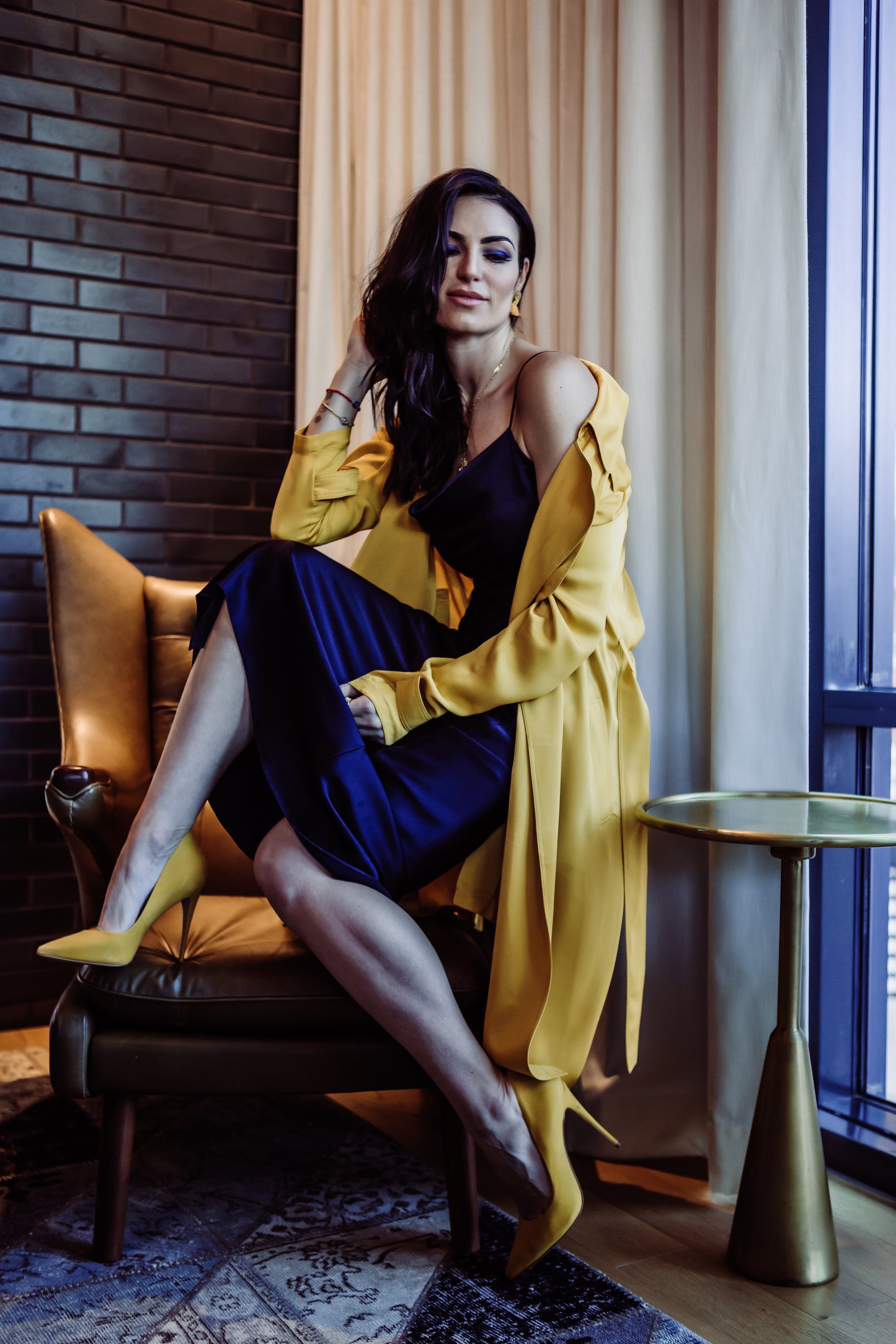 A woman sitting on a leather chair next to a window, wearing a black dress, yellow coat, and yellow high heels, with dark hair and makeup.