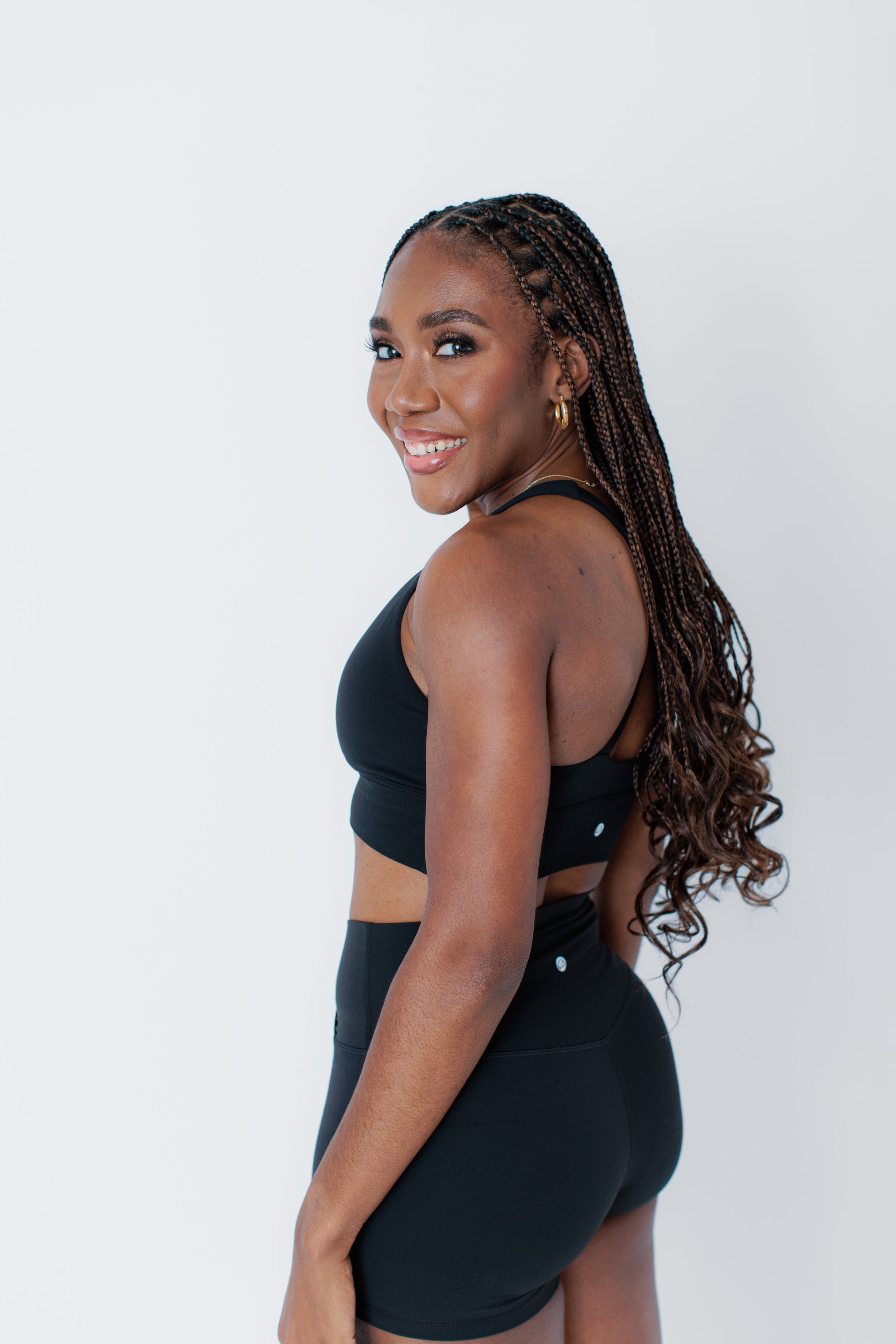 A young woman with long braided hair, smiling, wearing a black sports bra and matching high-waisted shorts, standing against a plain white background.