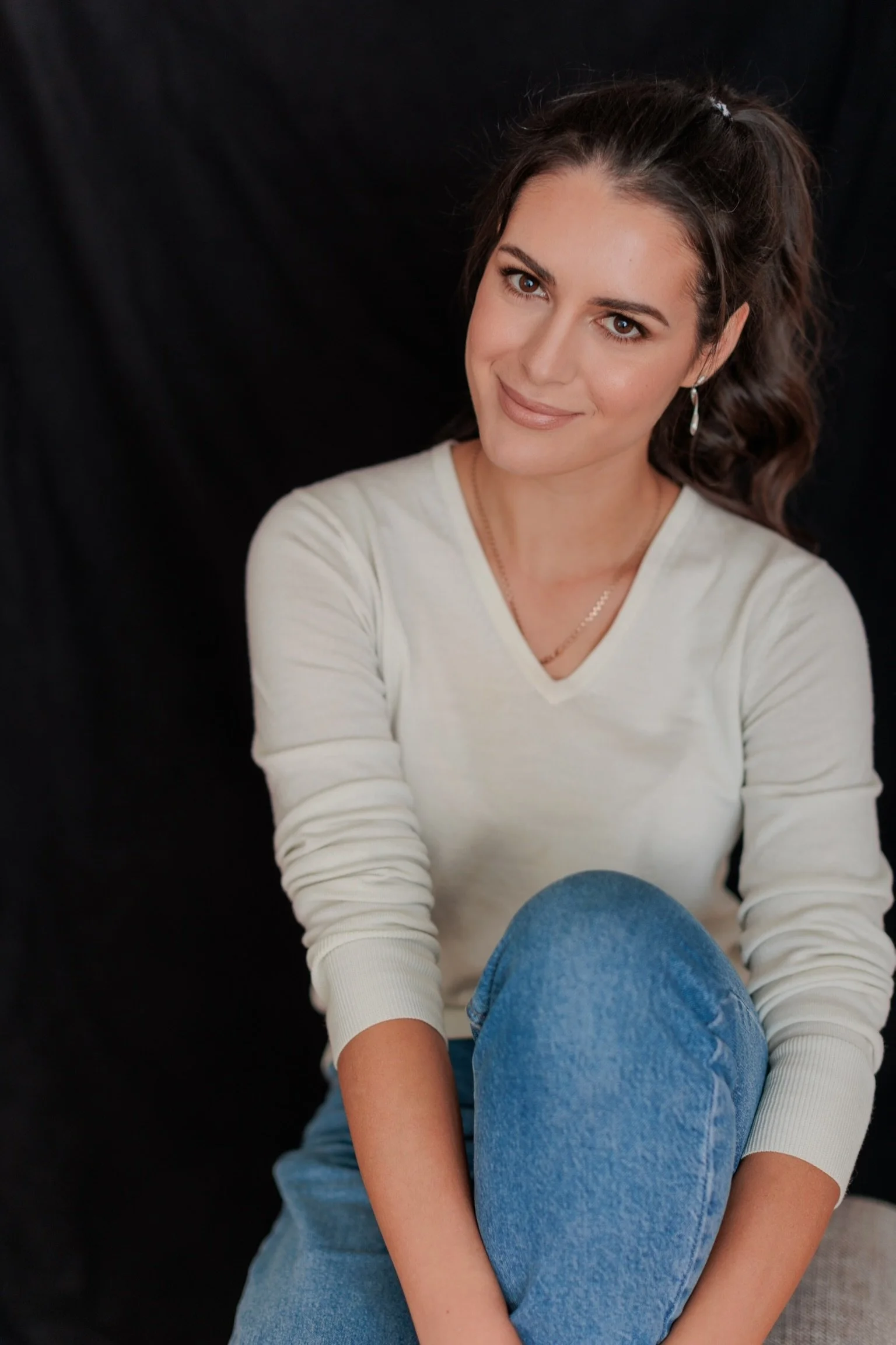 A young woman with long brown hair, wearing a white long sleeve shirt, blue jeans, and earrings, smiling at the camera against a black background.