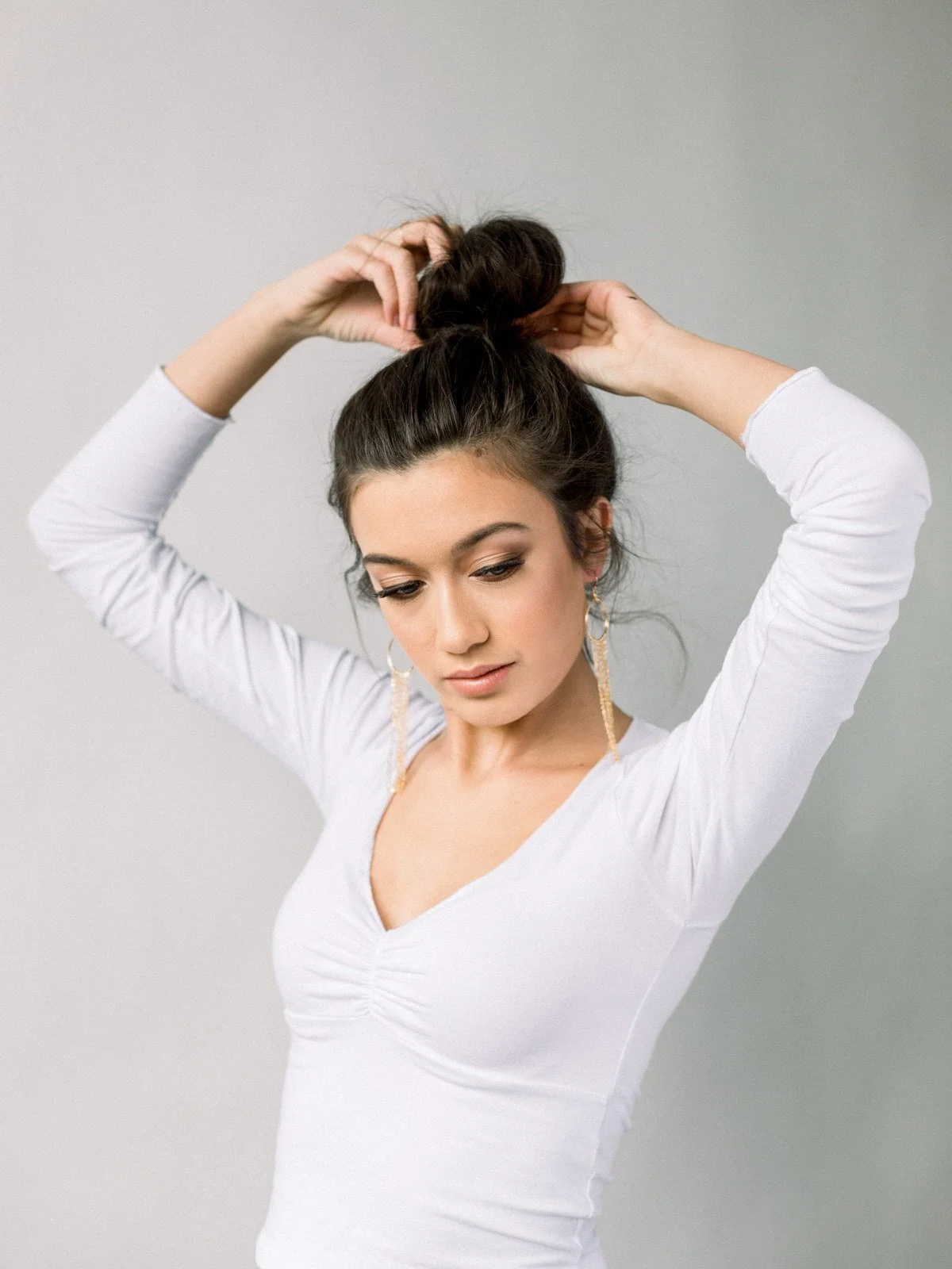 A young woman with dark hair styled in a high messy bun, wearing a white long-sleeve top and dangling earrings, standing against a plain background, adjusting her hair.