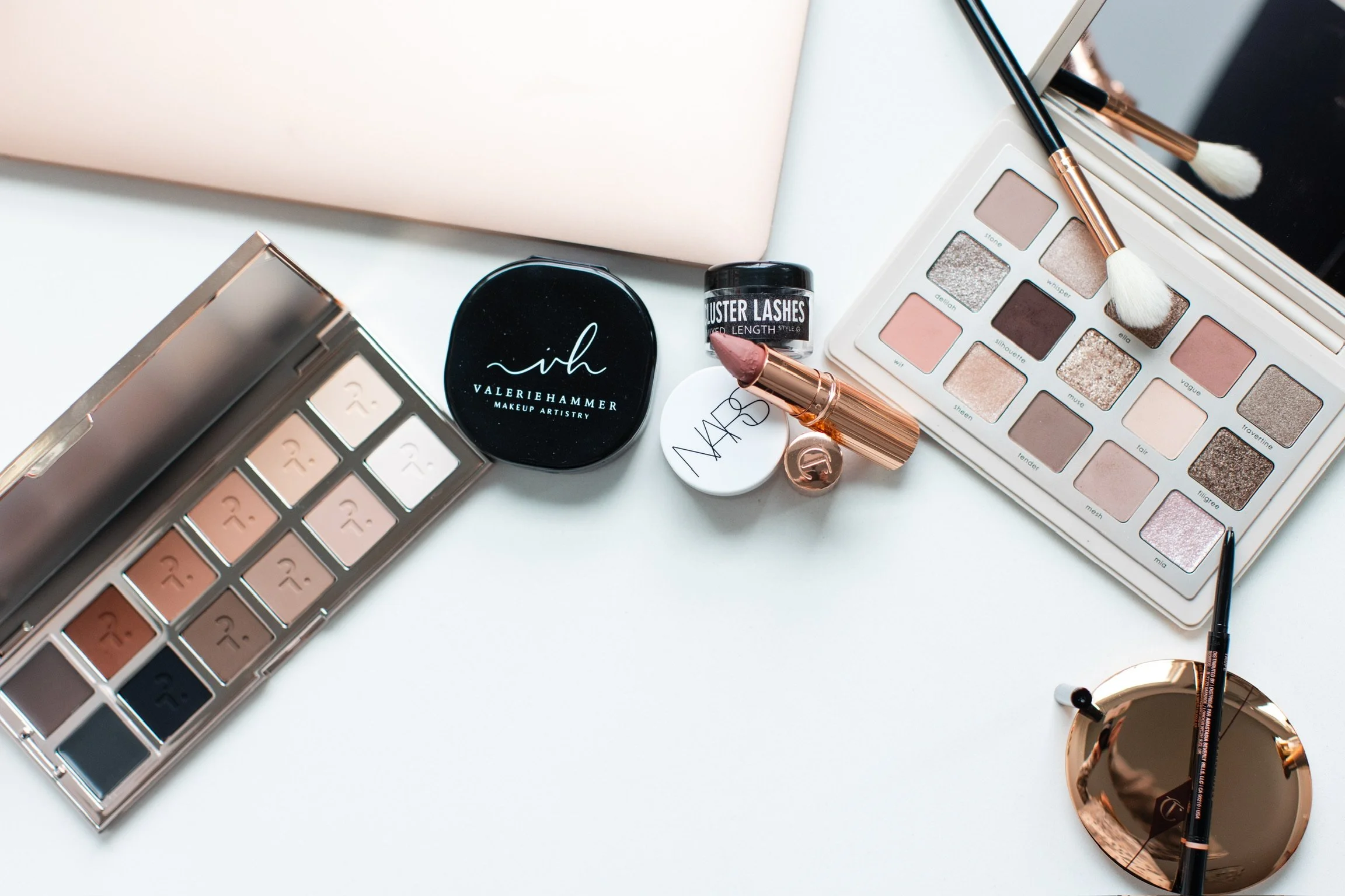 Flat lay of various makeup products on a white surface, including eyeshadow palettes, lipstick, mascara, brush, compact mirror, and skincare jars.