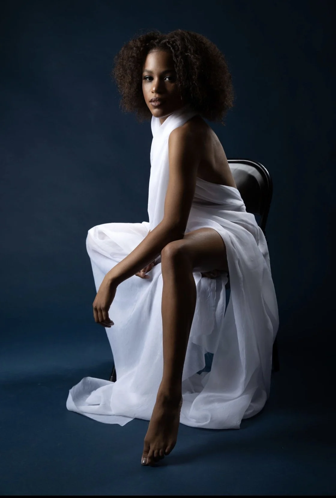 A woman with curly hair sitting on a chair, wrapped in a white fabric, against a dark background.