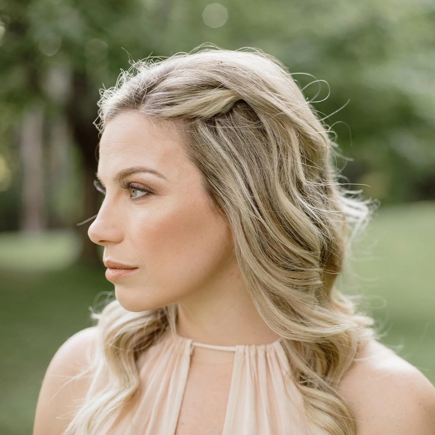 Profile of a woman with blonde, wavy hair outdoors, wearing a light-colored dress.