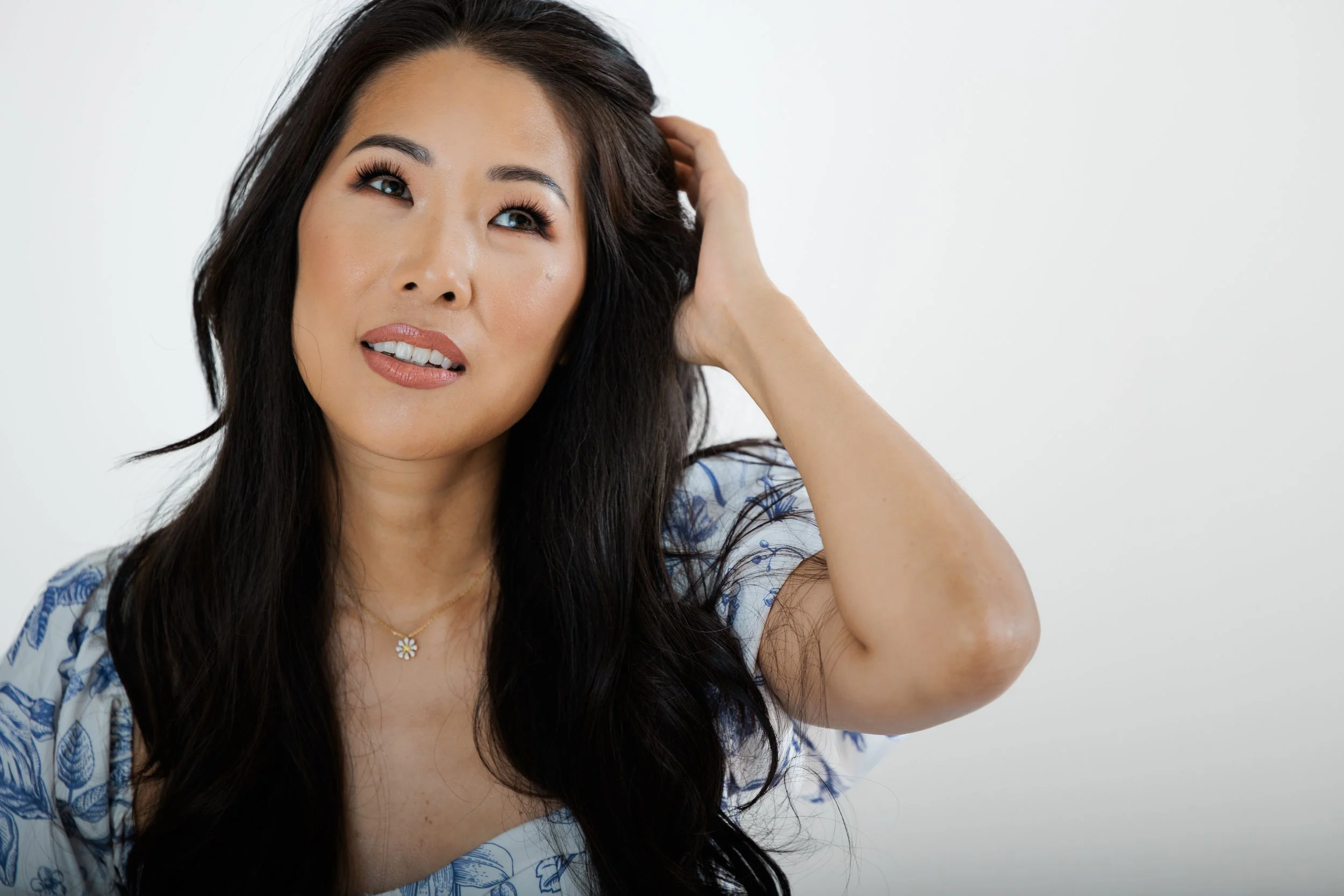 A woman with long dark hair, wearing a floral dress and a daisy necklace, touches her hair while looking upwards with a confused or puzzled expression against a plain white background.