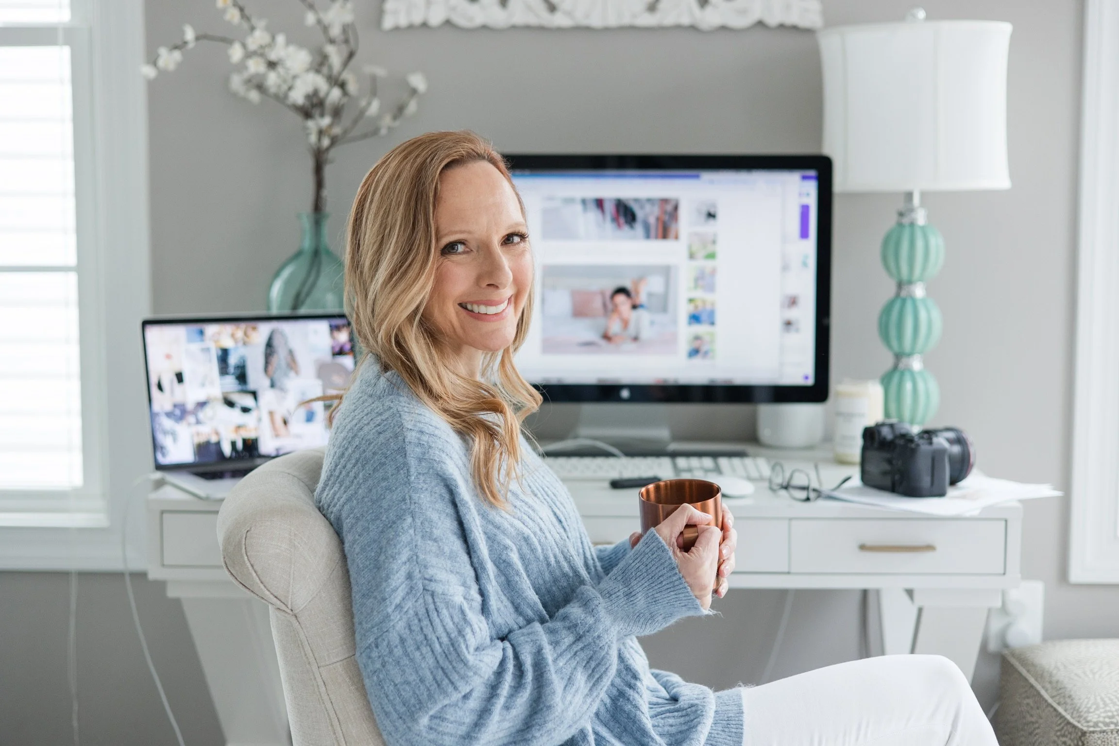 A woman sitting in a bright, modern home office, holding a coffee mug and smiling at the camera. Behind her are two computer monitors displaying photos and videos, along with a camera and other office supplies on the desk.